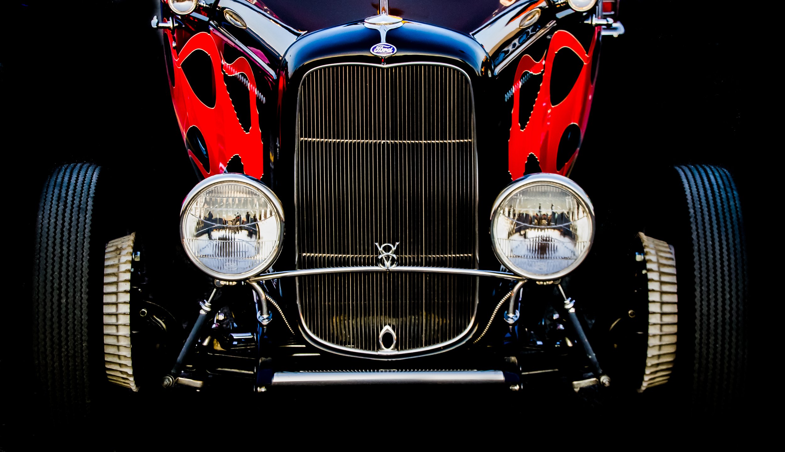 Front view of a vintage black and red Ford car with large headlights and a grille, reflecting people in a showroom or display area.