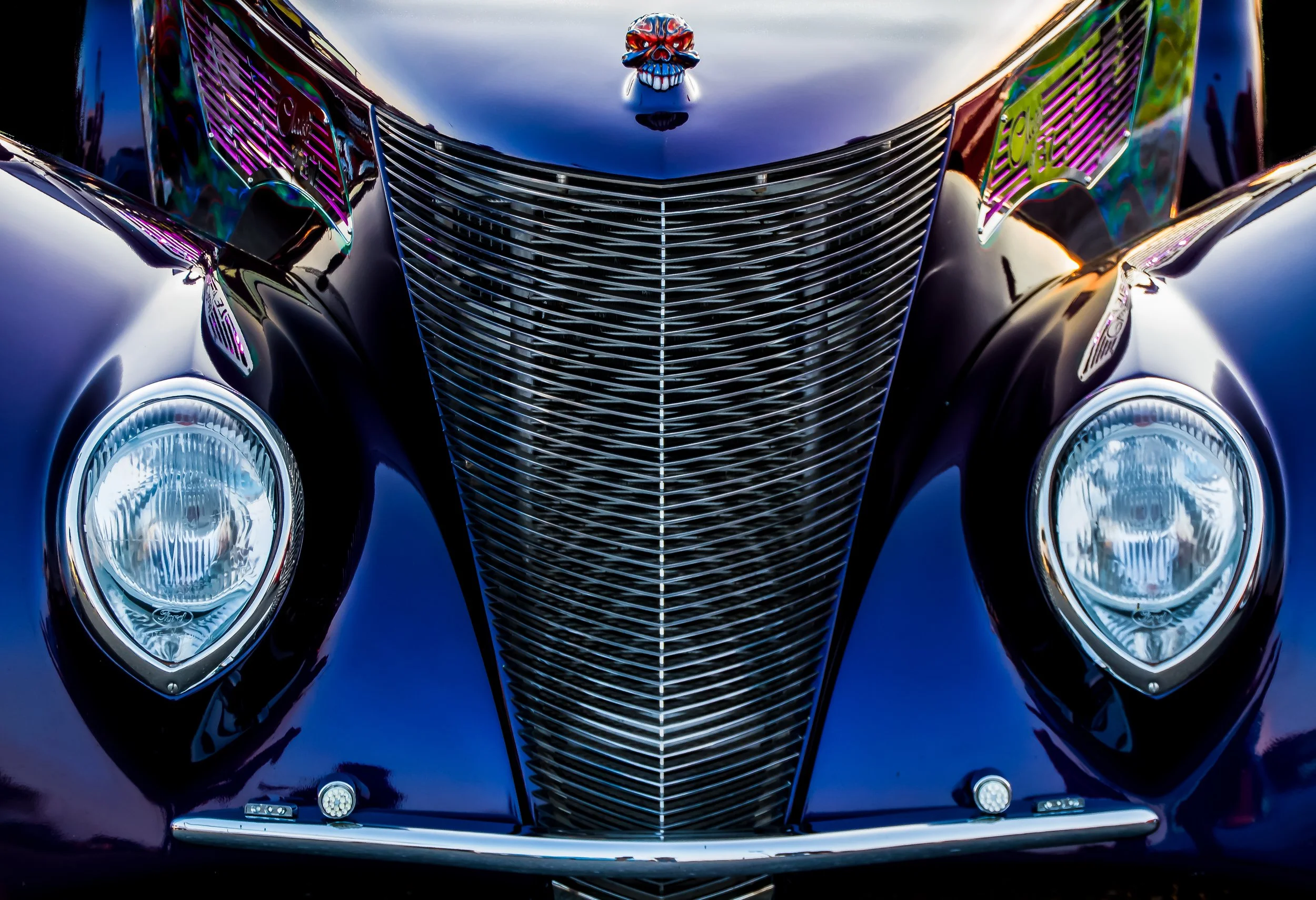 Front view of a shiny, vintage black car with a distinctive chrome grille, round headlights, and a skull ornament on the hood.