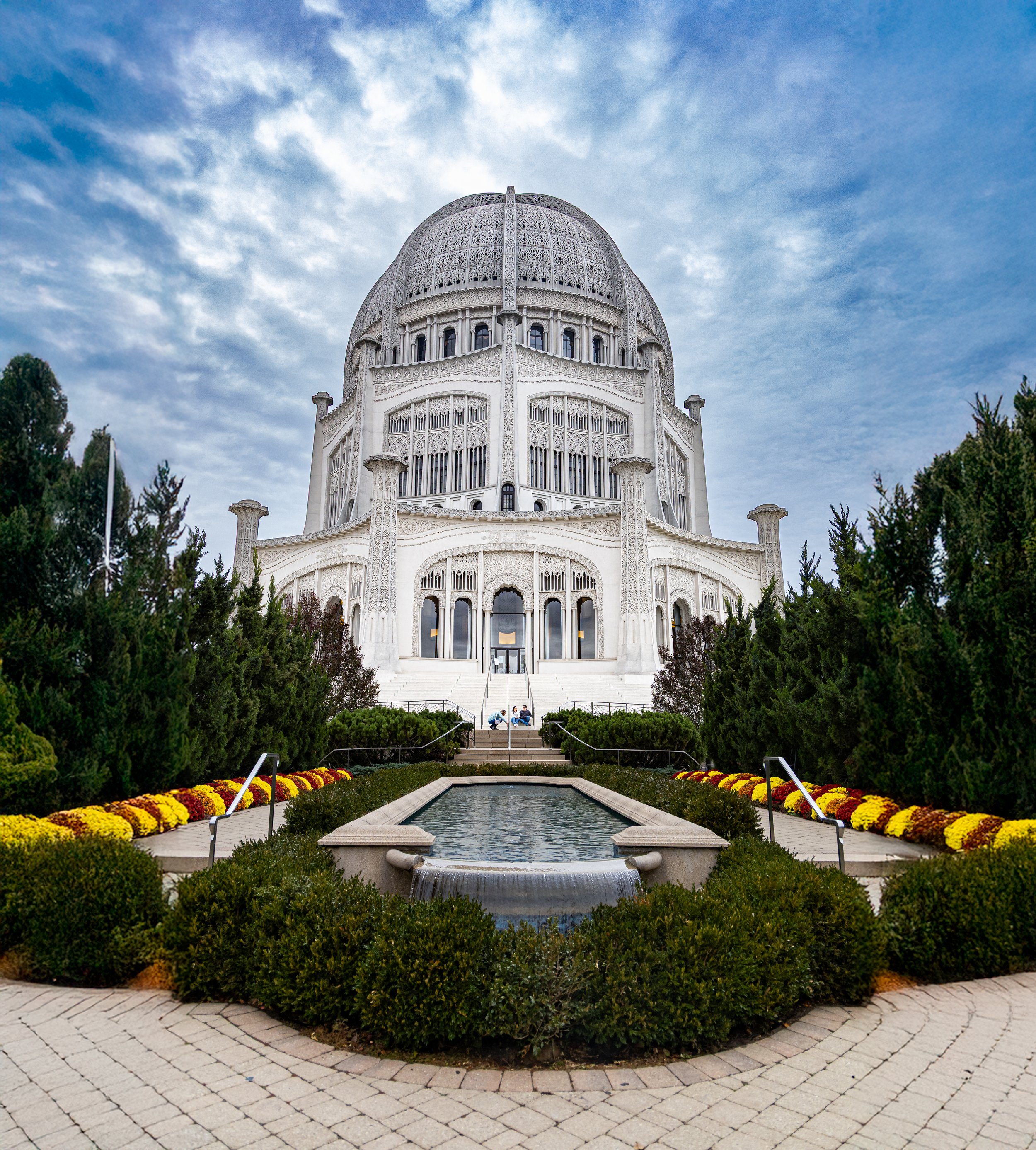 The Baha'i Temple in Wilmette, Illinois, featuring a white marble structure with intricate designs, a large central dome, surrounded by greenery, flower beds, and a reflecting pool in the foreground, under a partly cloudy sky.