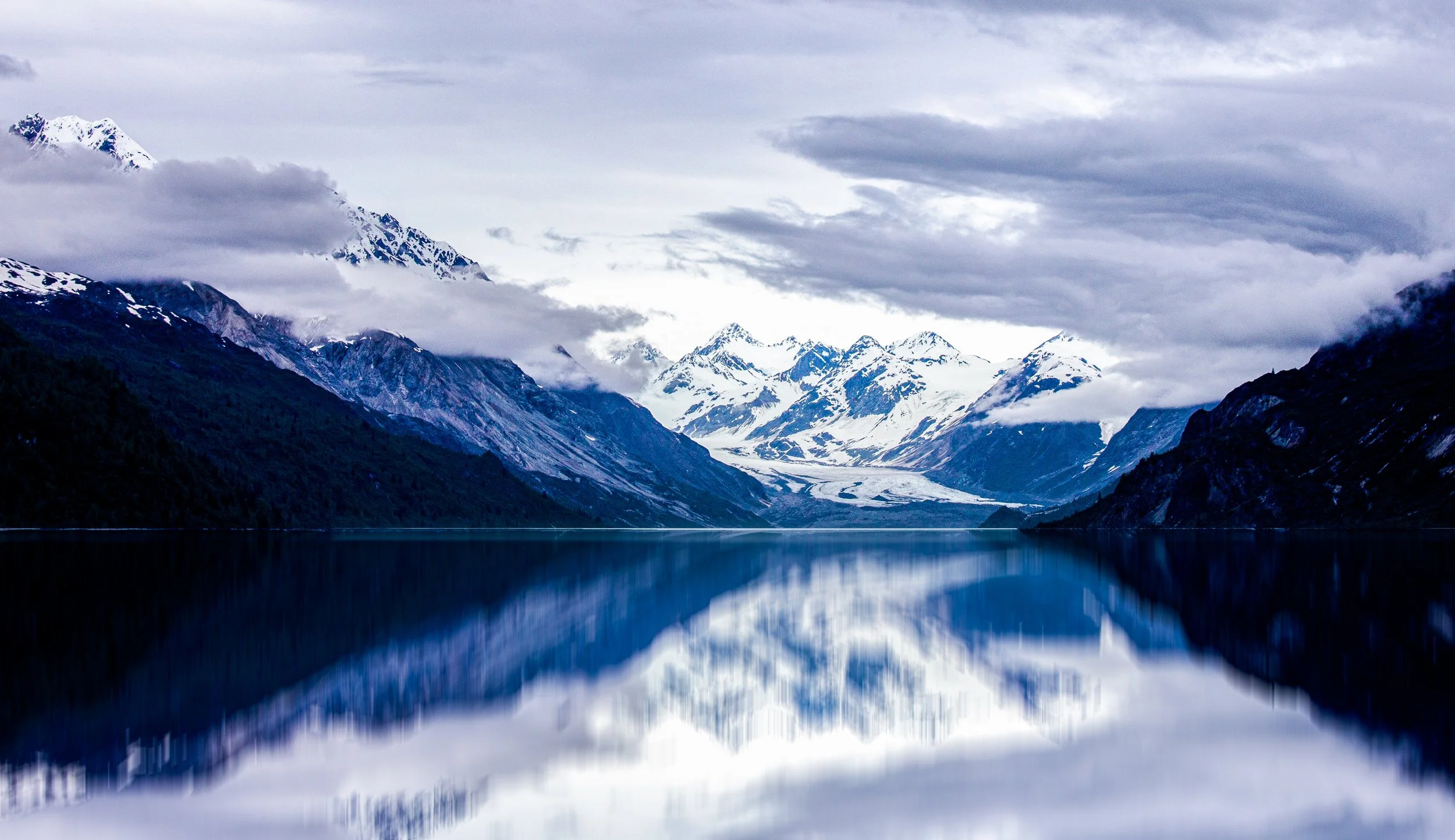 Mountain landscape with snow-capped peaks, cloudy sky, and a calm lake reflecting the mountains and sky.