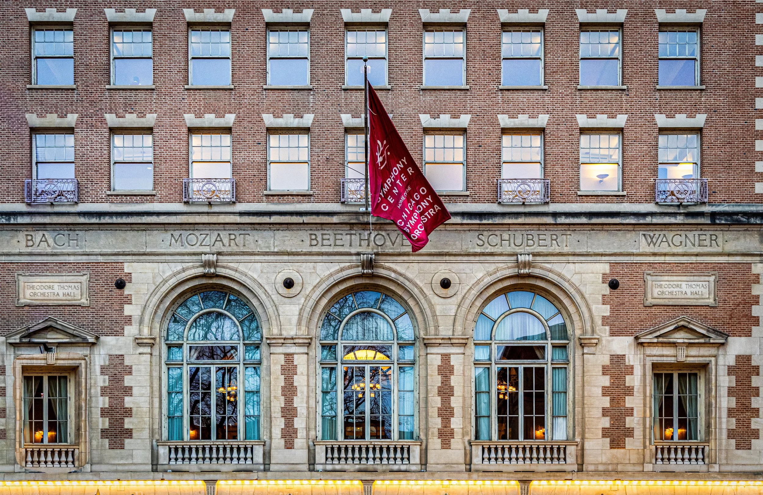 The facade of the Theodore Thomas Orchestra Hall, featuring large arched windows, a brick exterior, and a flag with the Chicago Symphony Orchestra logo hanging from the building.