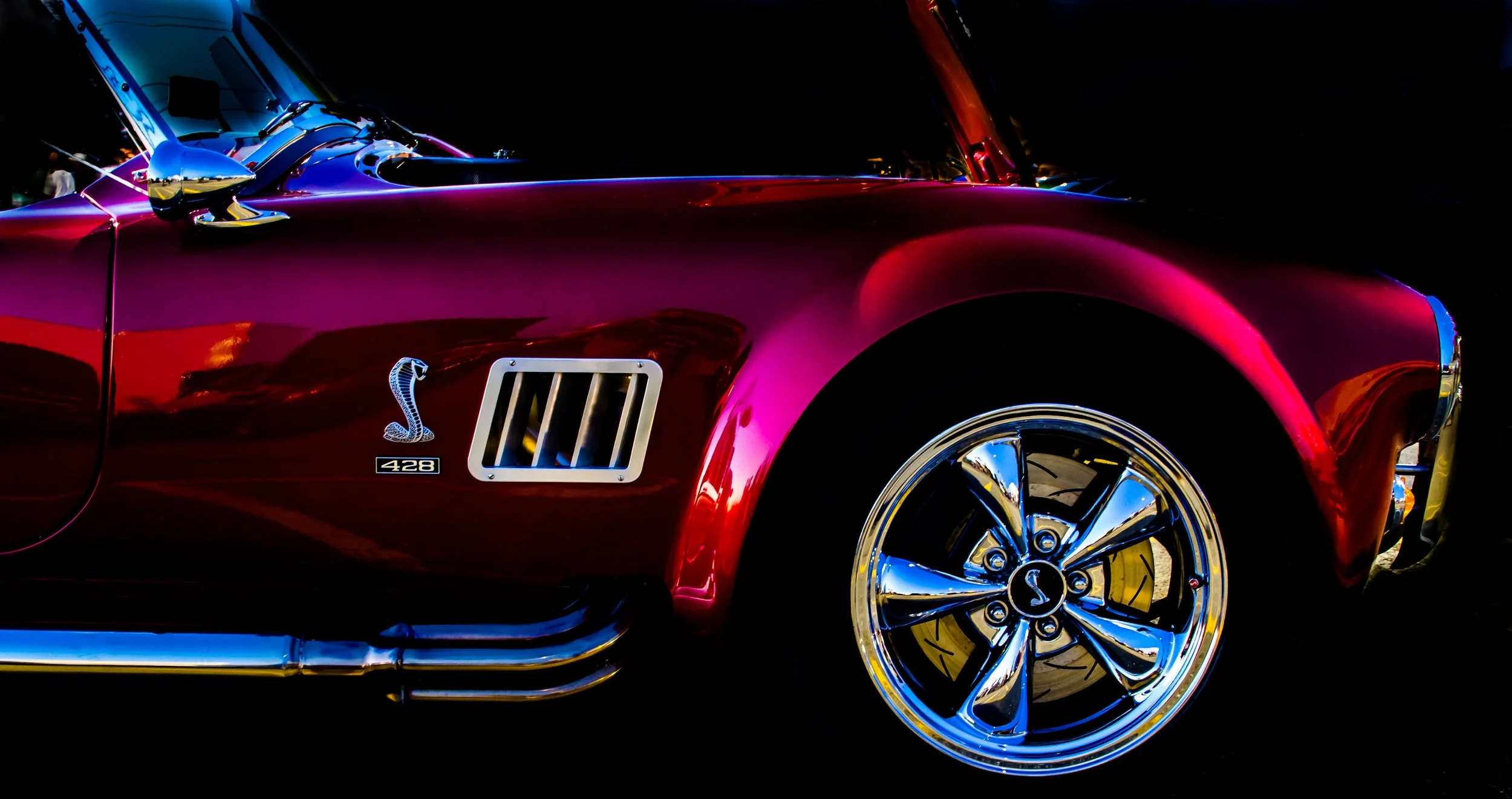 Close-up of a classic red Chevrolet Corvette with chrome accents, including a Cobra logo with the number 428 on the side, polished chrome wheel, and parts of the body and mirrors visible at night.
