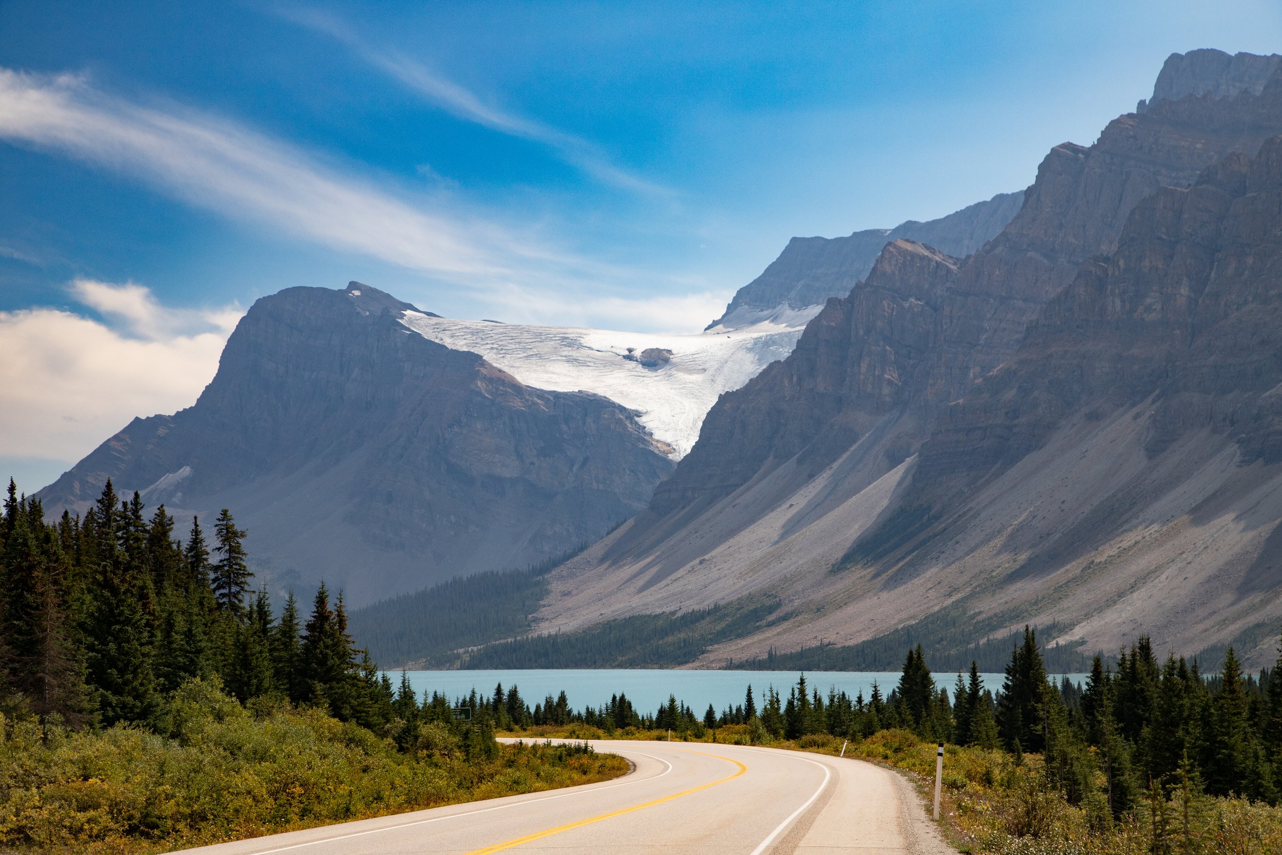 Scenic view of a winding road leading towards a turquoise lake, with tall pine trees and rugged mountains with glaciers in the background under a partly cloudy sky. Glacier. Rocky Mountain.  Ice parkways