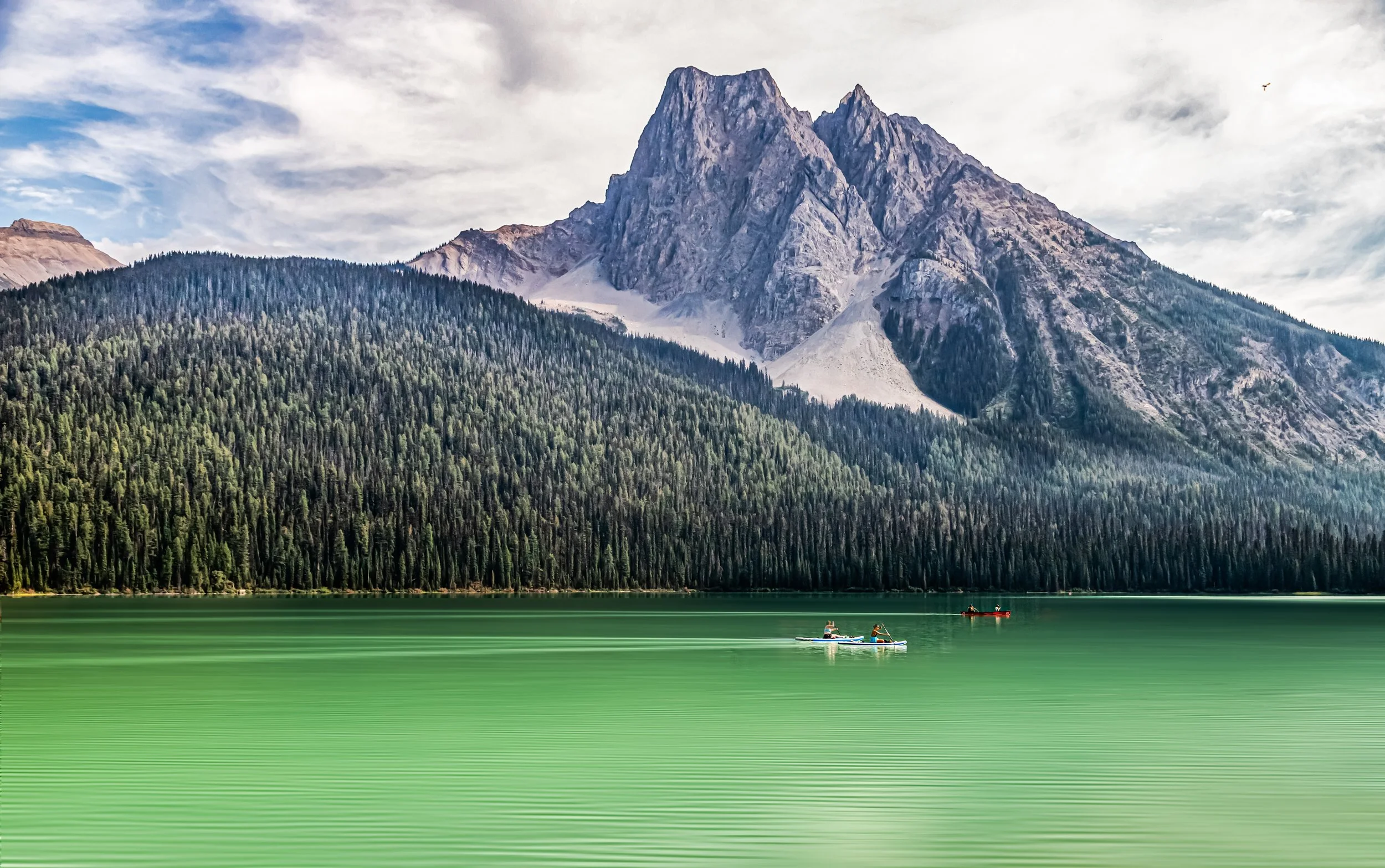 A scenic mountain landscape with a large mountain, dense green forest, and calm green lake with two small boats and two people paddling on the lake.