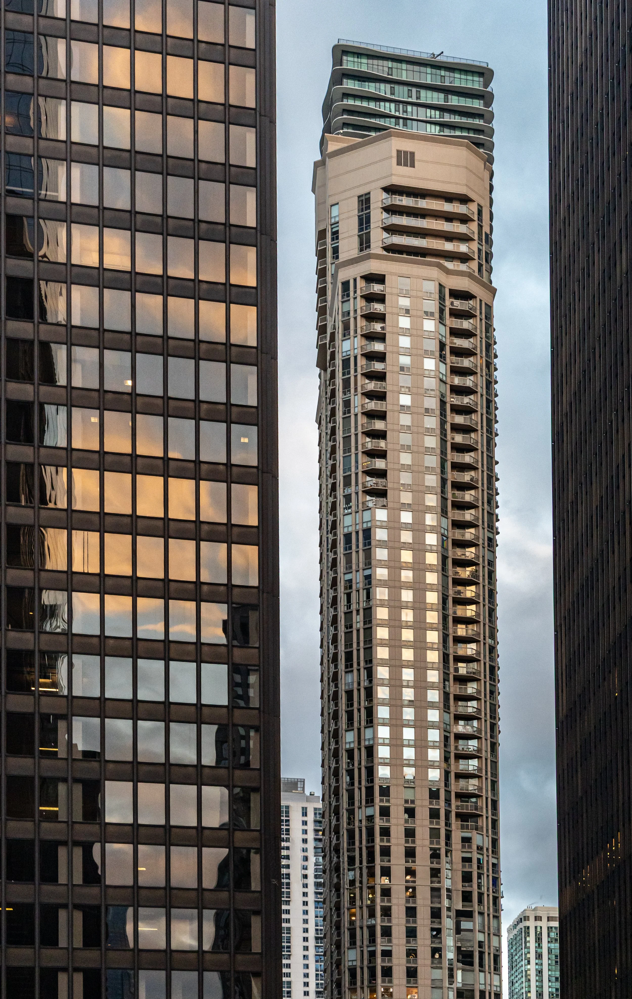 Cityscape with tall skyscrapers, including a modern high-rise with wavy balconies and a glass building reflecting the cloudy sky.