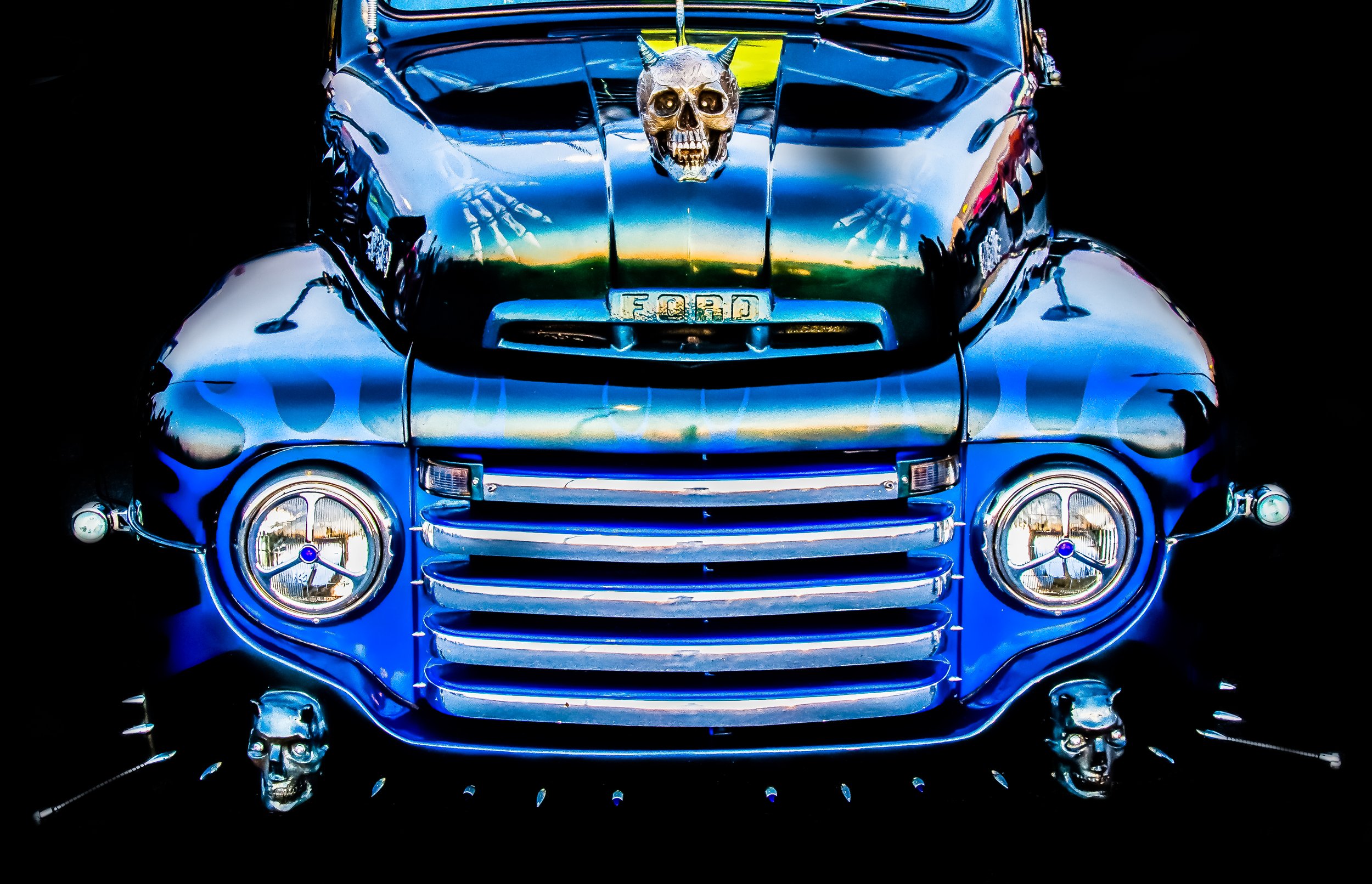 Front view of a shiny blue vintage car with skull and devil horn decoration on the hood, skull accents on the headlights, and skulls on the ground in front.
