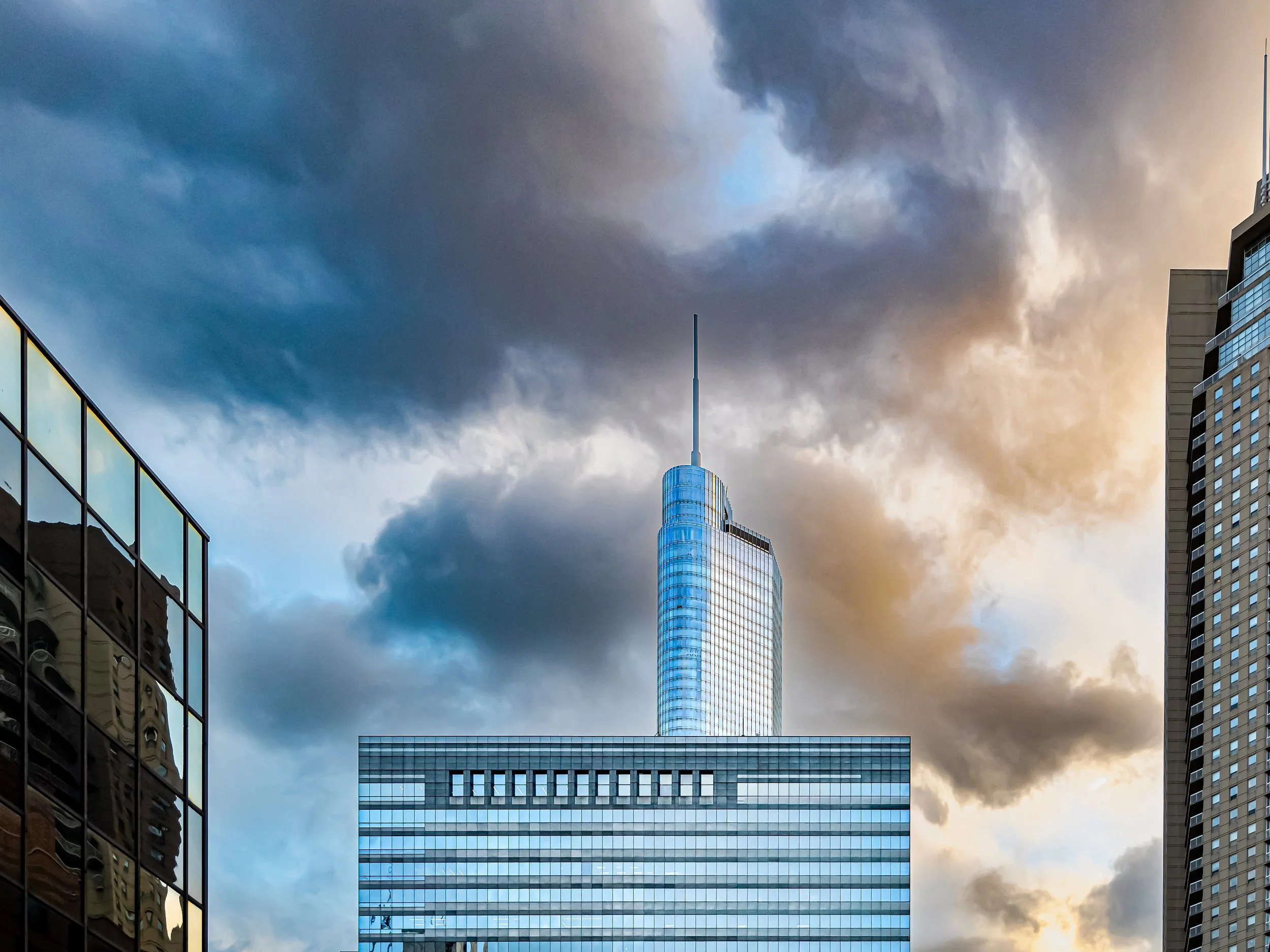 City skyline featuring a tall glass building with a spire, alongside other skyscrapers, under a dramatic sky with dark and light clouds.