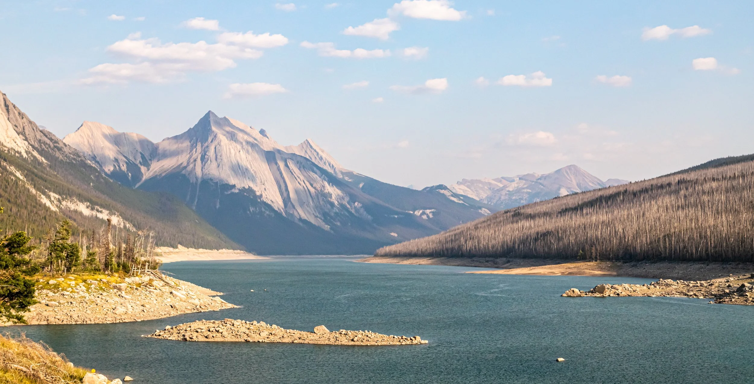 Scenic view of a mountain lake with calm waters, surrounded by mountain ranges and partially forested hills under a partly cloudy sky.