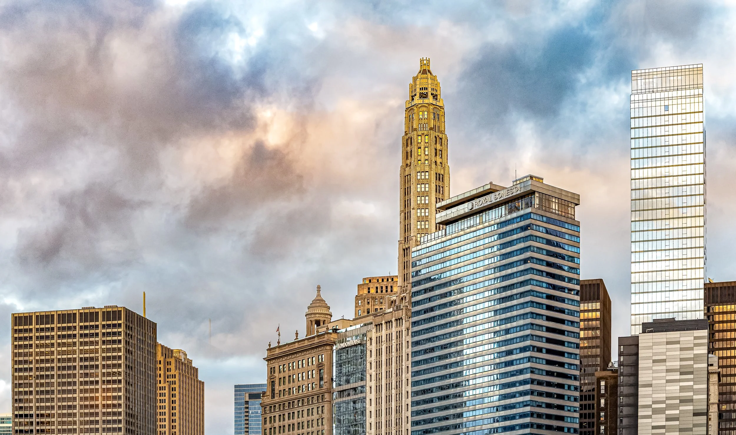 City skyline with tall skyscrapers pointing towards a cloudy sky, including a prominent golden Art Deco tower and modern glass buildings.