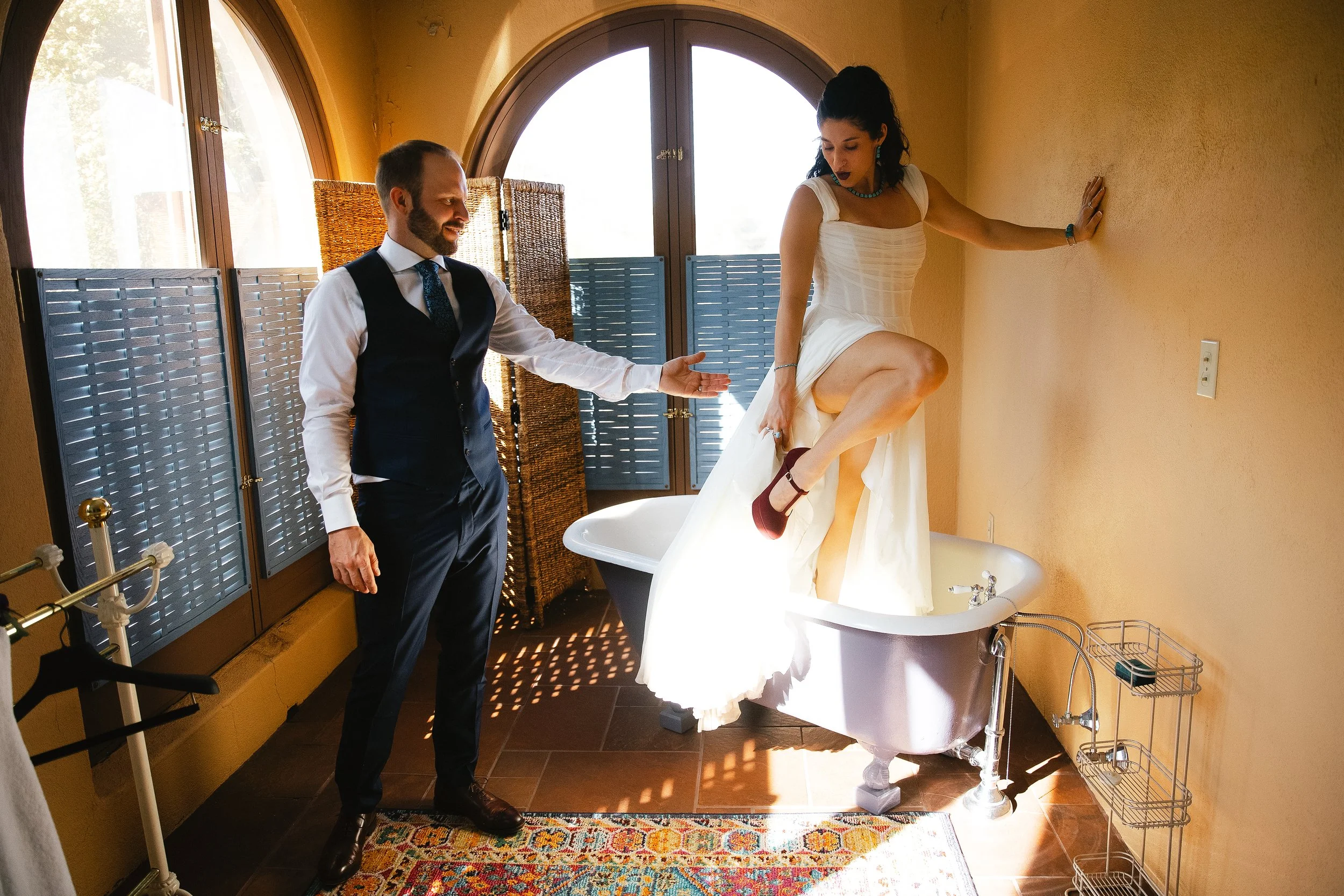 A bride in a white dress stepping into a bathtub with assistance from the groom in a suit and vest, inside a brightly lit room with arched windows and yellow walls at the Little Daisy, a wedding venue in Jerome, Arizona. Photographed by Megan Farmer,