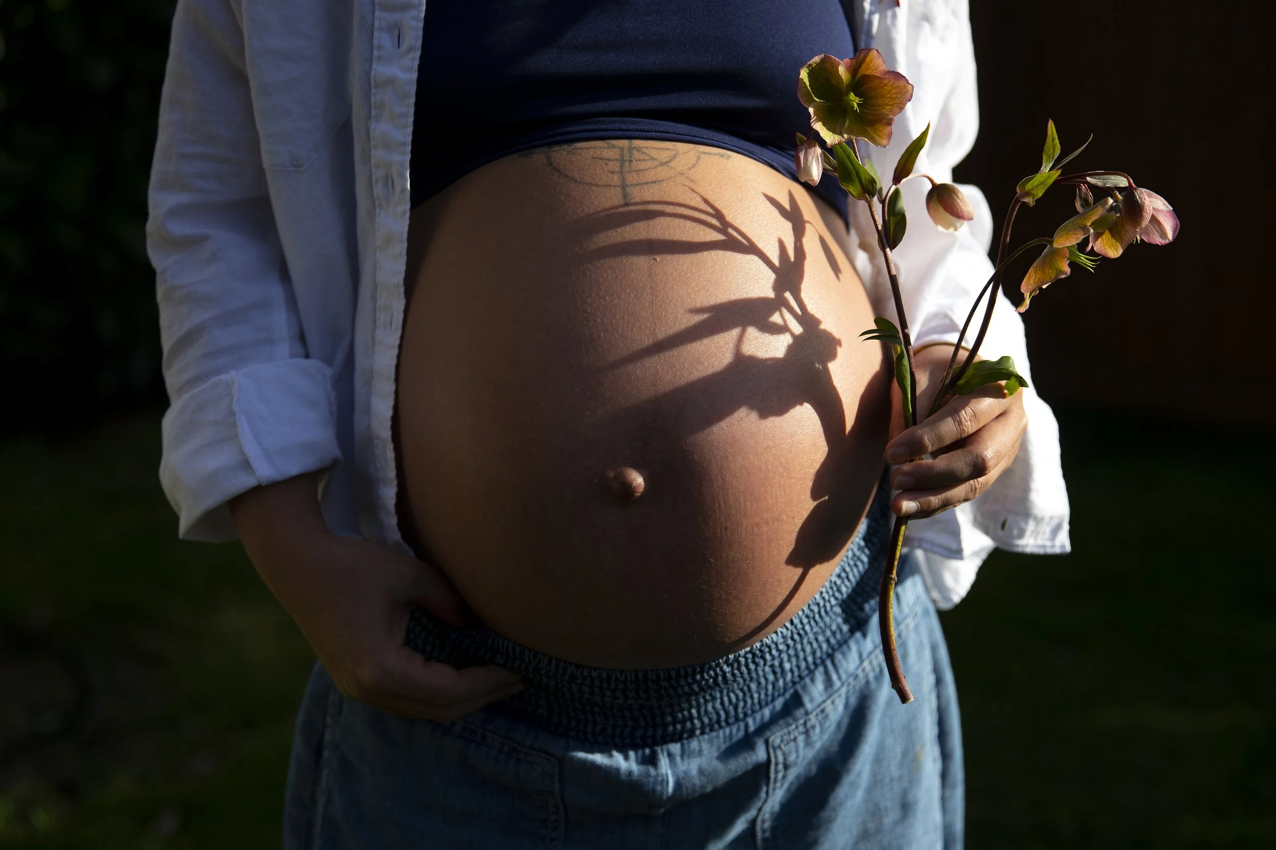 A pregnant woman holding pink flowers, with the shadow of the flowers cast on her stomach while being photographed in North Bend, Washington, by Megan Farmer, a documentary style wedding and family photographed based in Seattle, Washington. 