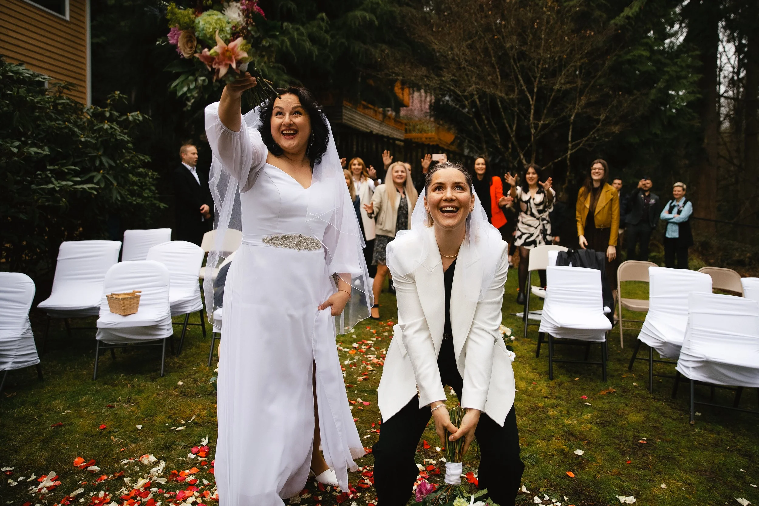 Two women, one in a white wedding dress and the other in a white blazer and black pants, prepare to throw their wedding bouquets while celebrating outdoors with friends and family following their wedding ceremony. Guests are smiling and cheering in t