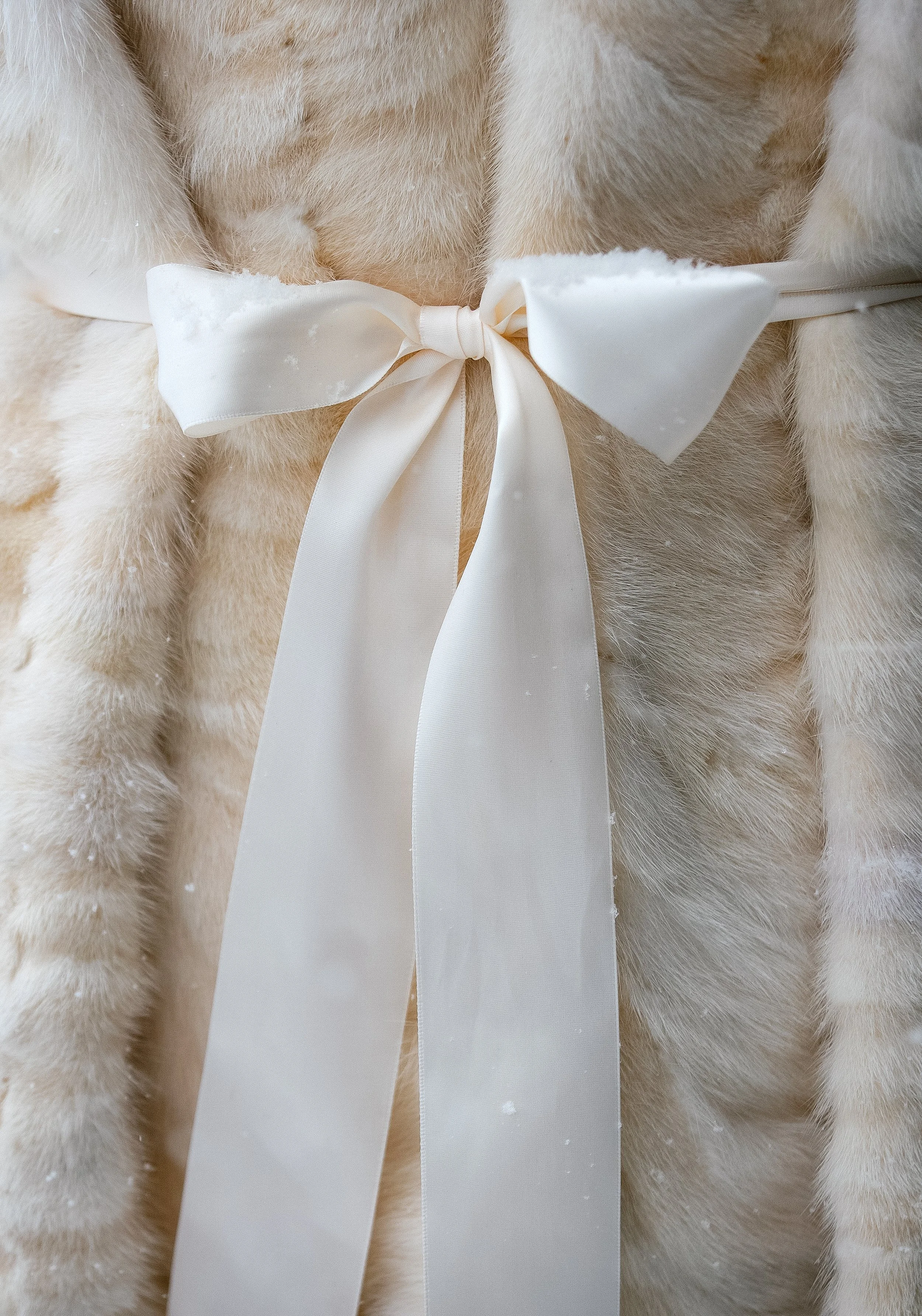 Close-up of a fluffy beige fur coat with a satin white ribbon tied into a bow at a wedding ceremony photographed by documentary style wedding photographer Megan Farmer, at Golden Creek Pond in Snoqualmie Pass, Washington.
