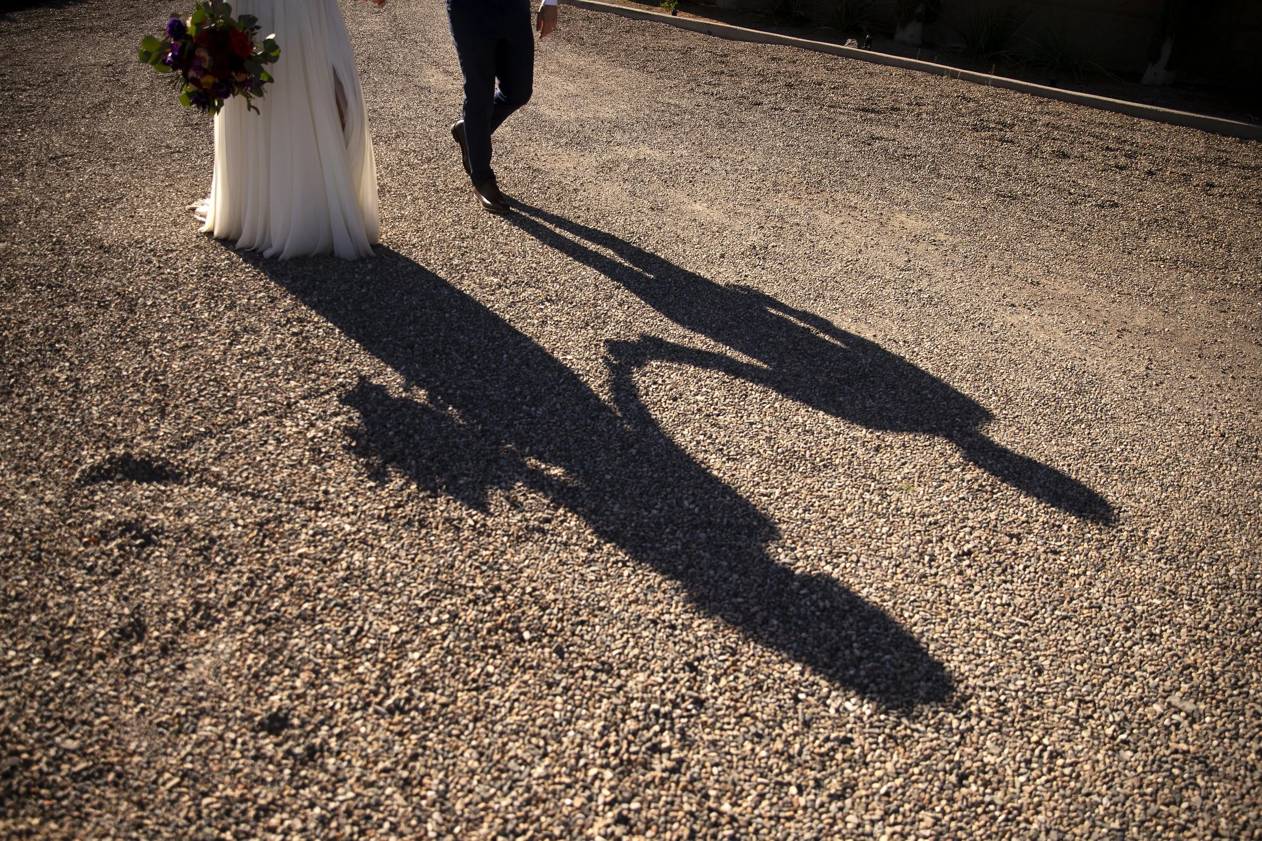 Bride and groom walking on gravel, with their long shadows cast on the ground, one holding a bouquet of flowers, before their wedding ceremony at the Little Daisy, a venue in Jerome, Arizona. Photographed by Megan Farmer, a documentary style wedding 
