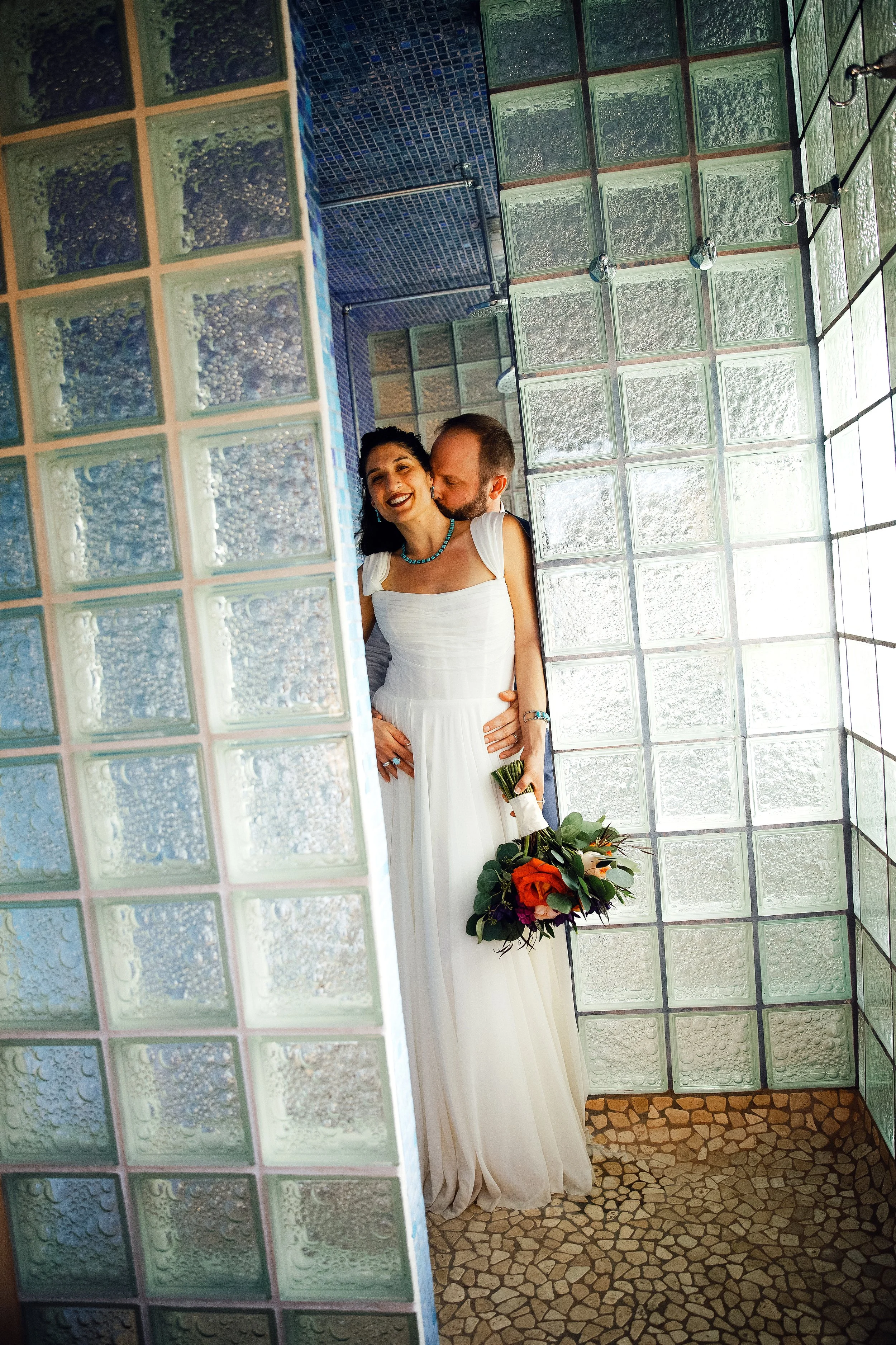 A bride and groom pose for a portrait before their wedding ceremony, standing in a shower area with glass block walls at the Little Daisy, a wedding venue in Jerome, Arizona. Photographed by Megan Farmer, a documentary style wedding photographer base