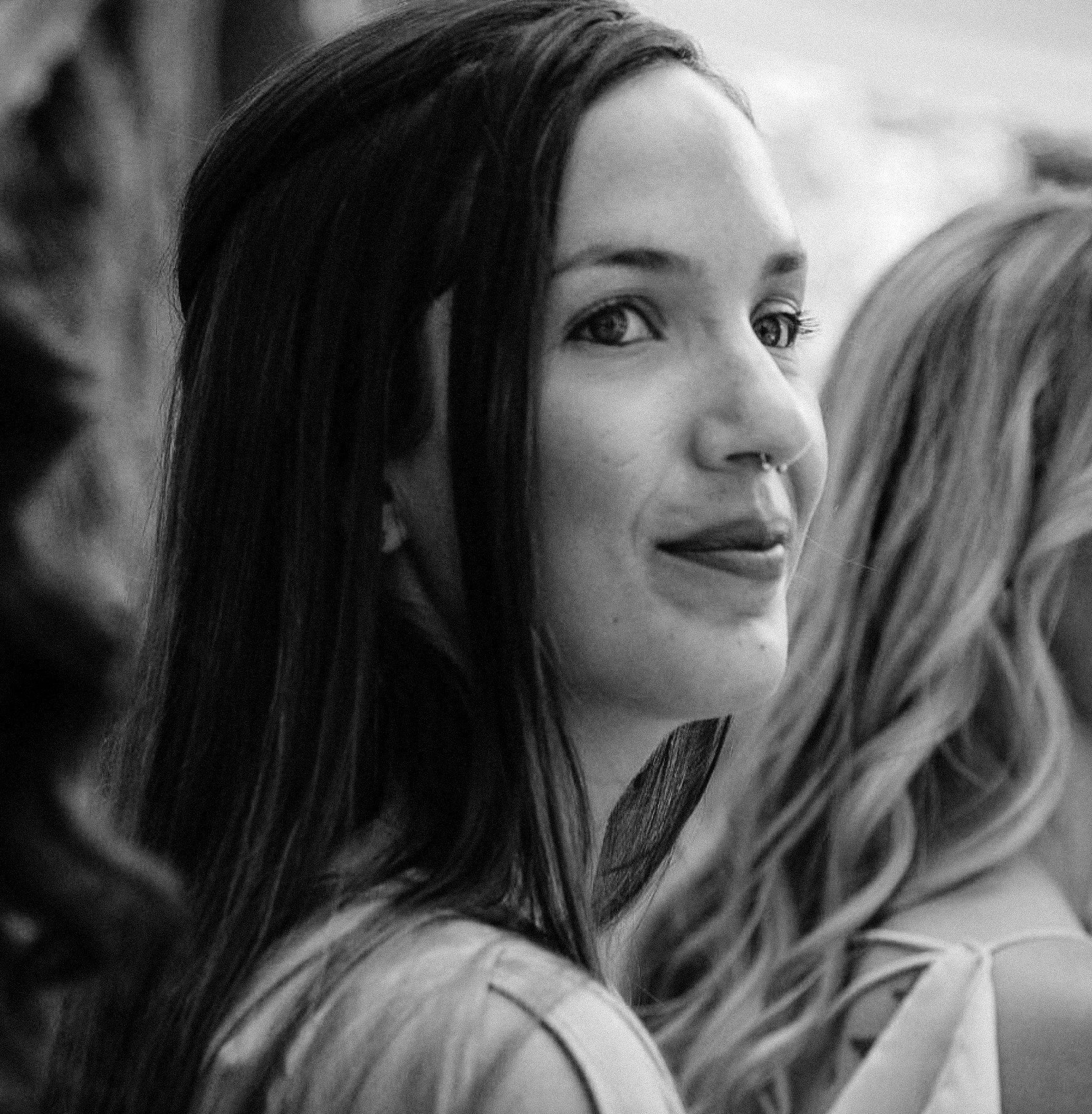 A young woman with long dark hair, septum piercing, and earrings, looking over her shoulder at the camera, with a slight smile. She is flanked by other women, one with curly hair on the right and another obscured on the left.