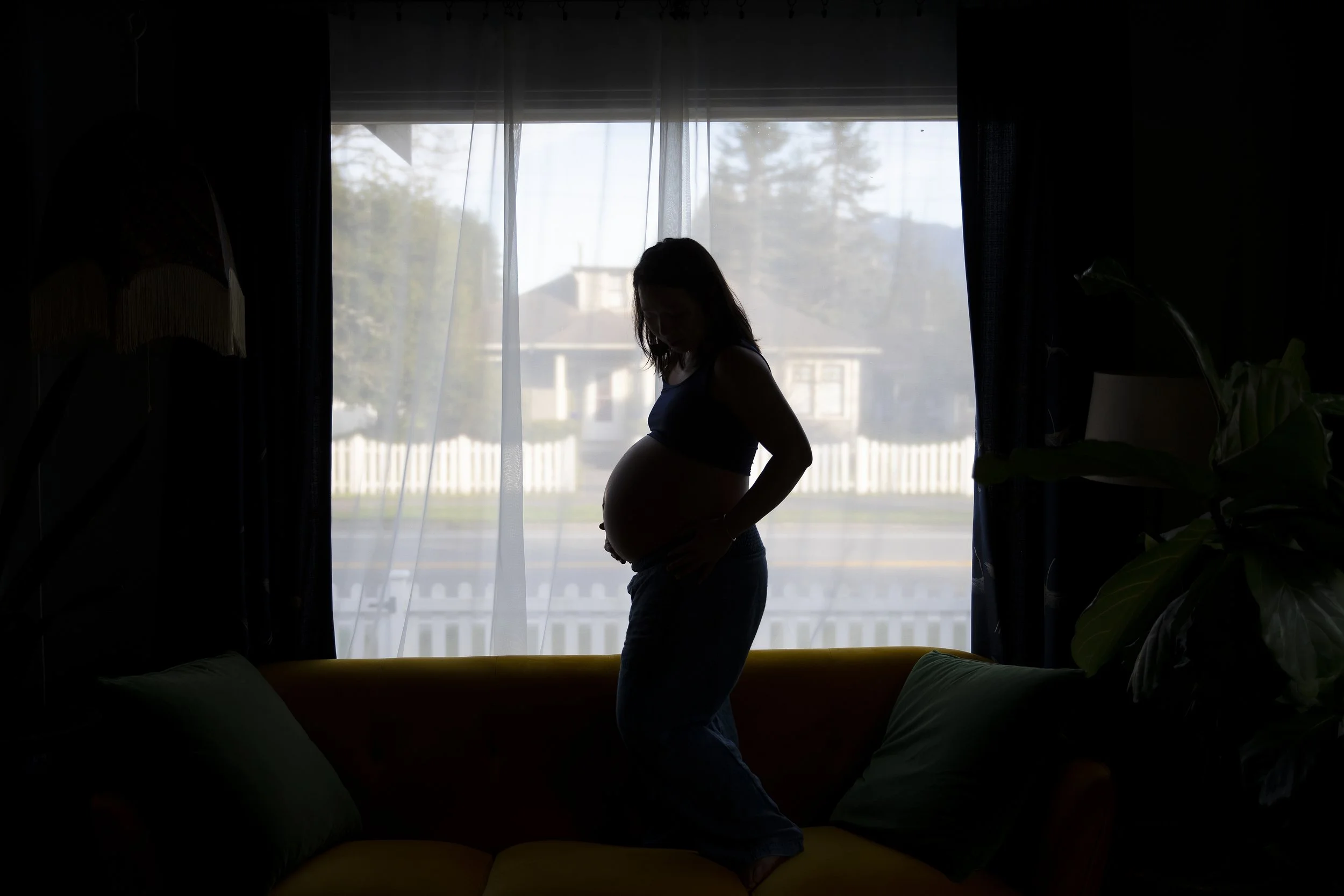 Silhouette of a pregnant woman standing on a couch in front of a window, with a suburban neighborhood visible outside, in North Bend, Washington, photographed by Megan Farmer, a documentary style wedding and family photographer based in Seattle, Wash