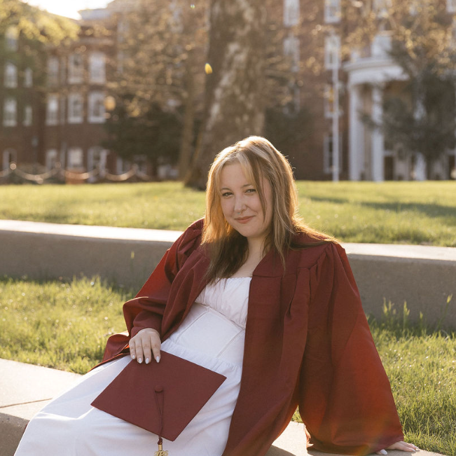 A young woman in a graduation gown sitting on a sidewalk in a park, holding a graduation cap, with a residential building in the background, during sunset.