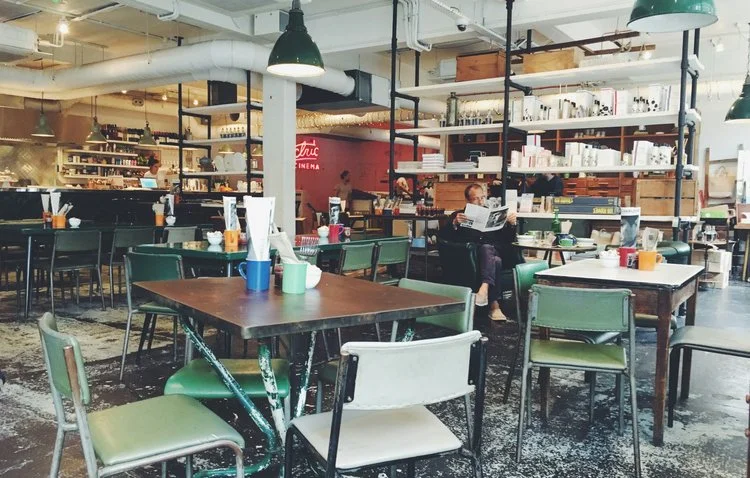 Interior of a cafe with tables and chairs, shelves with various items, and people sitting and reading; industrial decor with open ceiling and hanging lights.