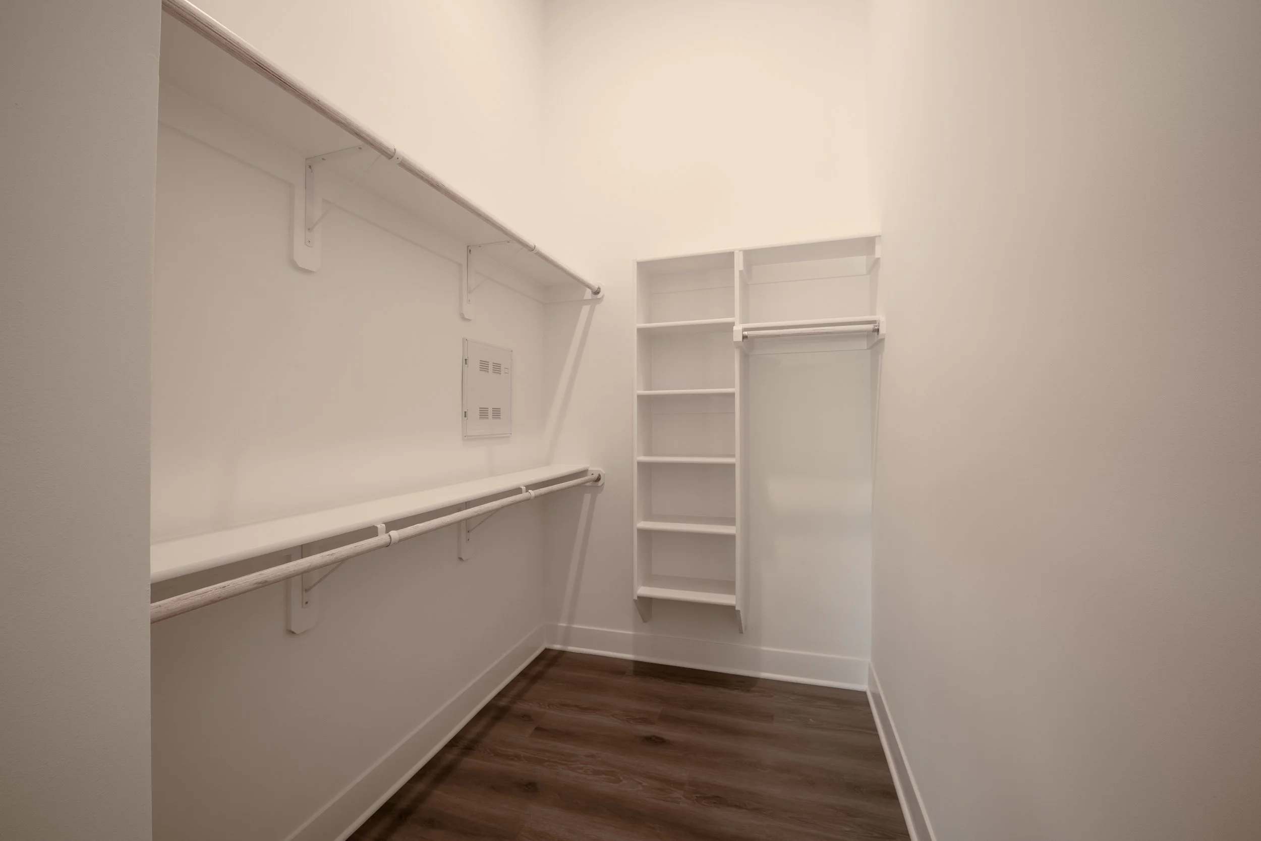 Empty closet with white shelves, rods for hanging clothes, and a brown hardwood floor.