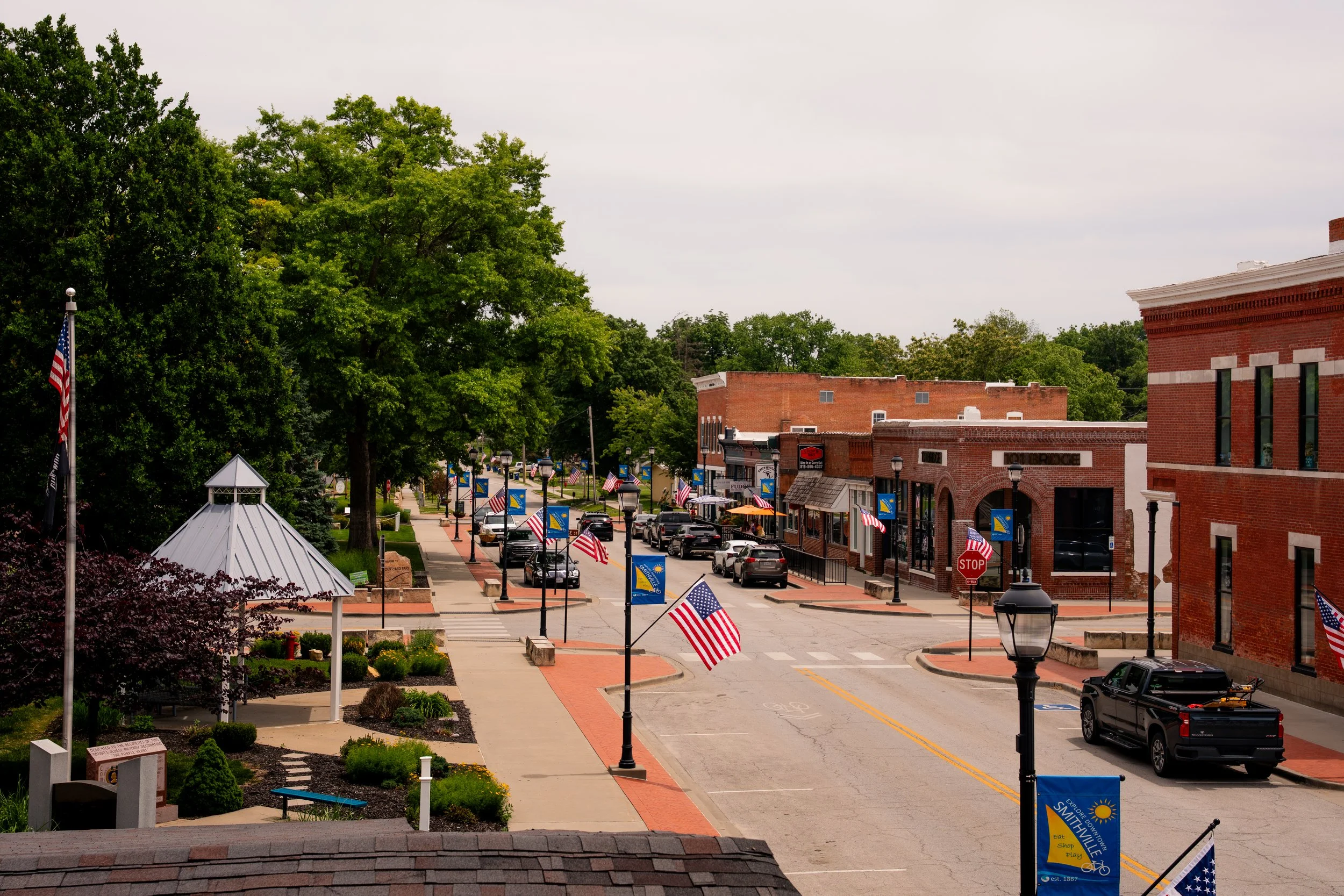 A small town main street decorated with American flags, trees on the sidewalk, brick buildings, parked cars, and street lamps, under a cloudy sky.