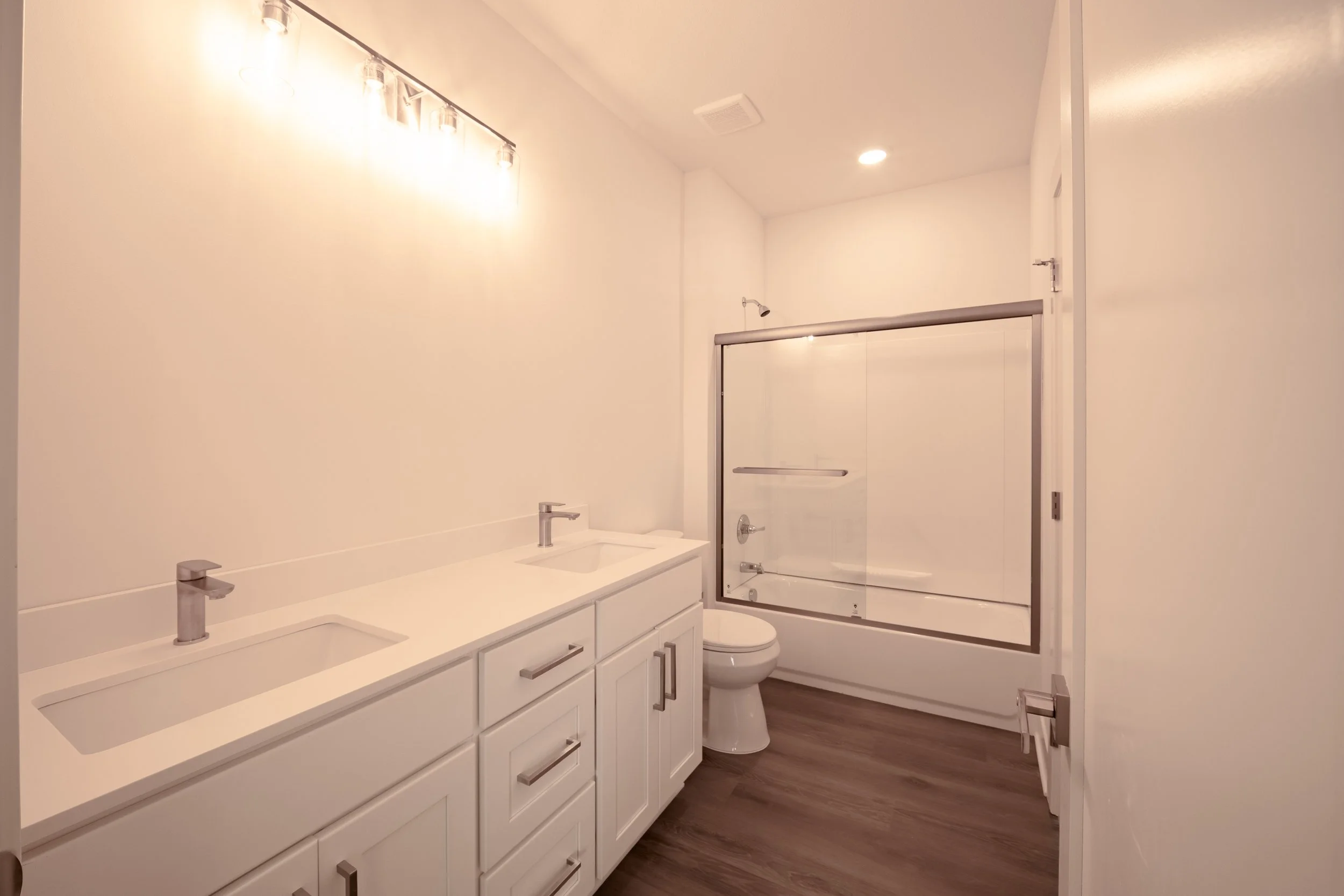 A modern bathroom with a double sink vanity, white cabinets, a toilet, a bathtub with a glass sliding door, and a showerhead. The walls are plain, and the ceiling has recessed lighting. The floor is wood or wood-like material.