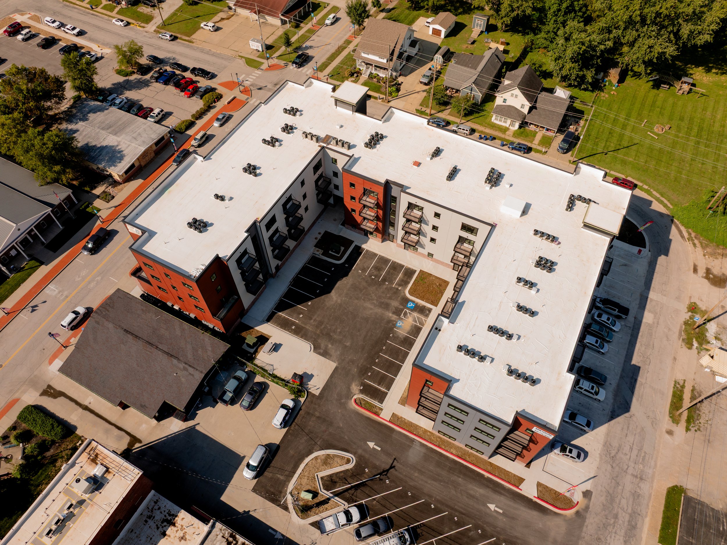 An aerial view of a multi-story apartment building featuring a large parking lot and air conditioning units. Surrounding the building are residential houses, a parking lot with cars, sidewalks, and green grassy areas with trees.