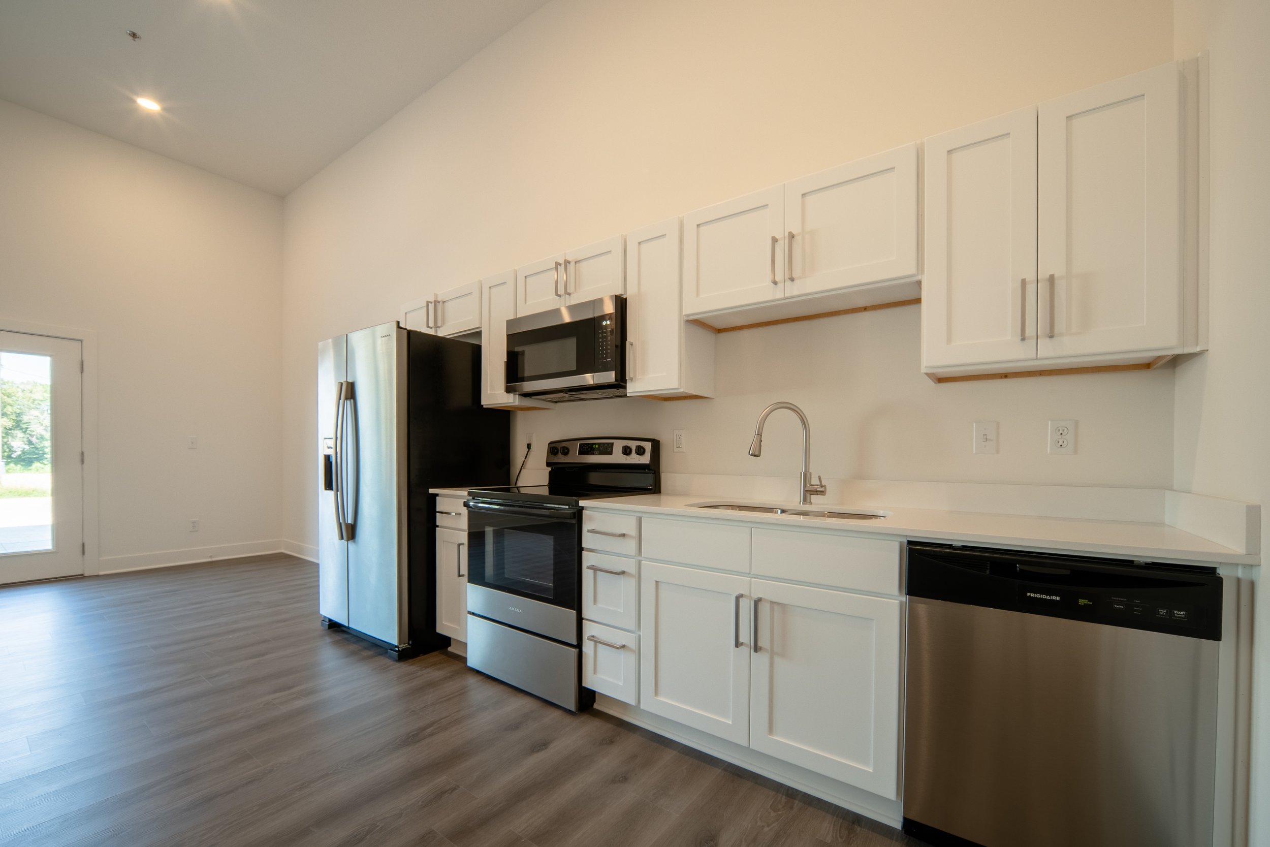 Modern kitchen with white cabinets, stainless steel refrigerator, black microwave, and stove, with wood-look flooring and a door with a window leading outside.