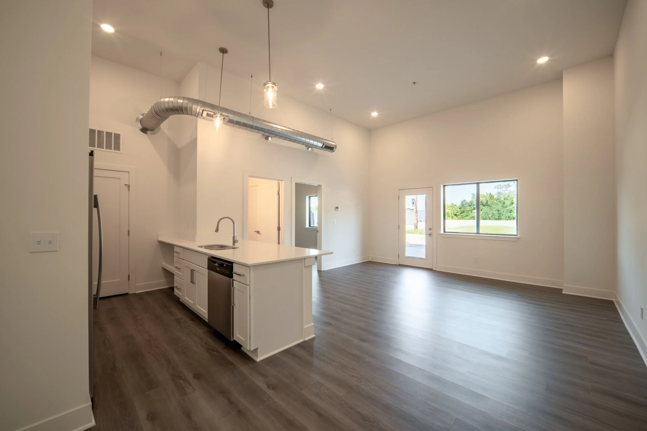 Empty modern kitchen and living area with large windows and high ceiling with exposed ductwork.