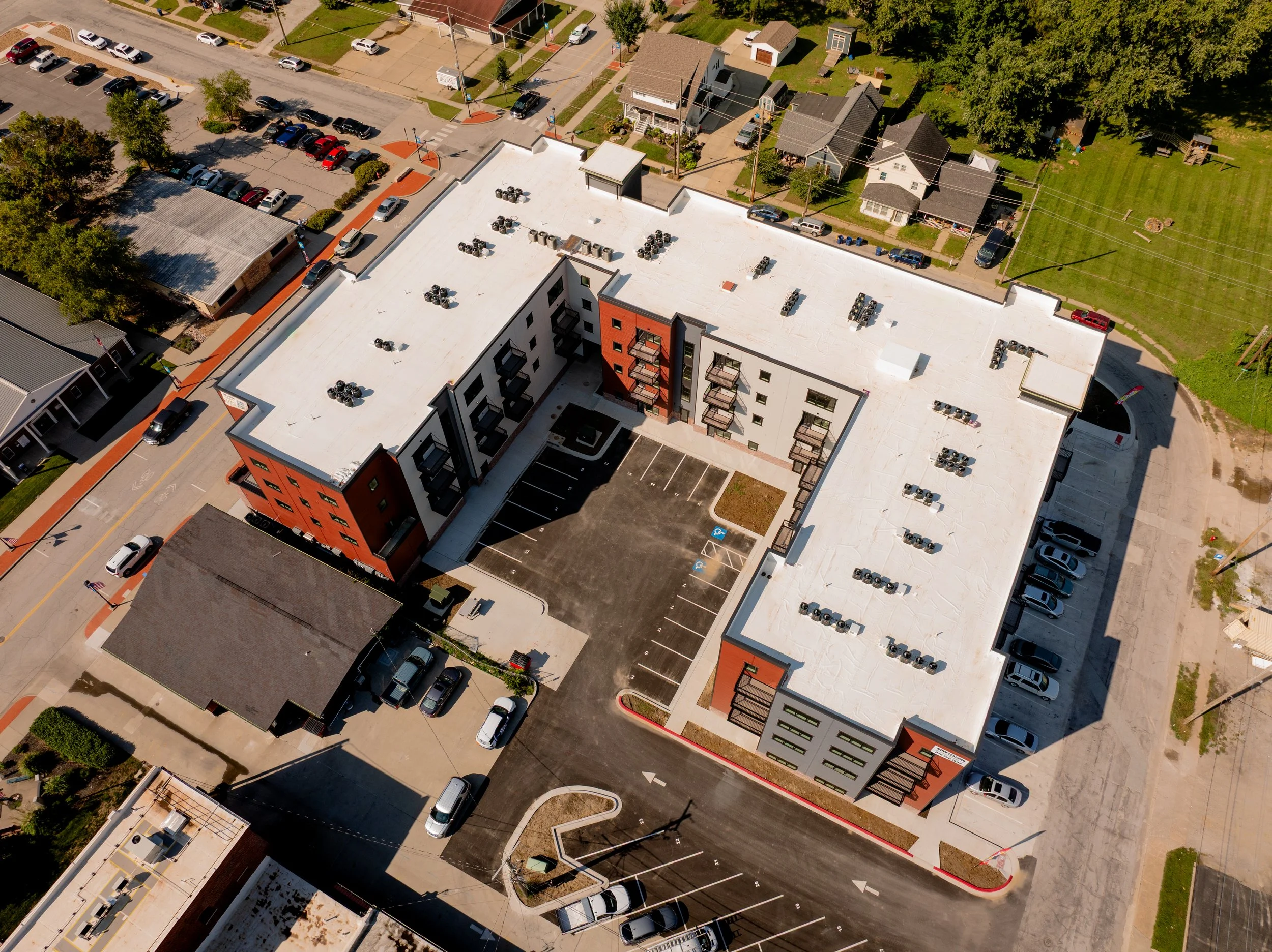 An aerial view of a multi-story apartment building featuring a large parking lot, air conditioning units, adjacent parking lot, surrounding residential homes, and green trees.