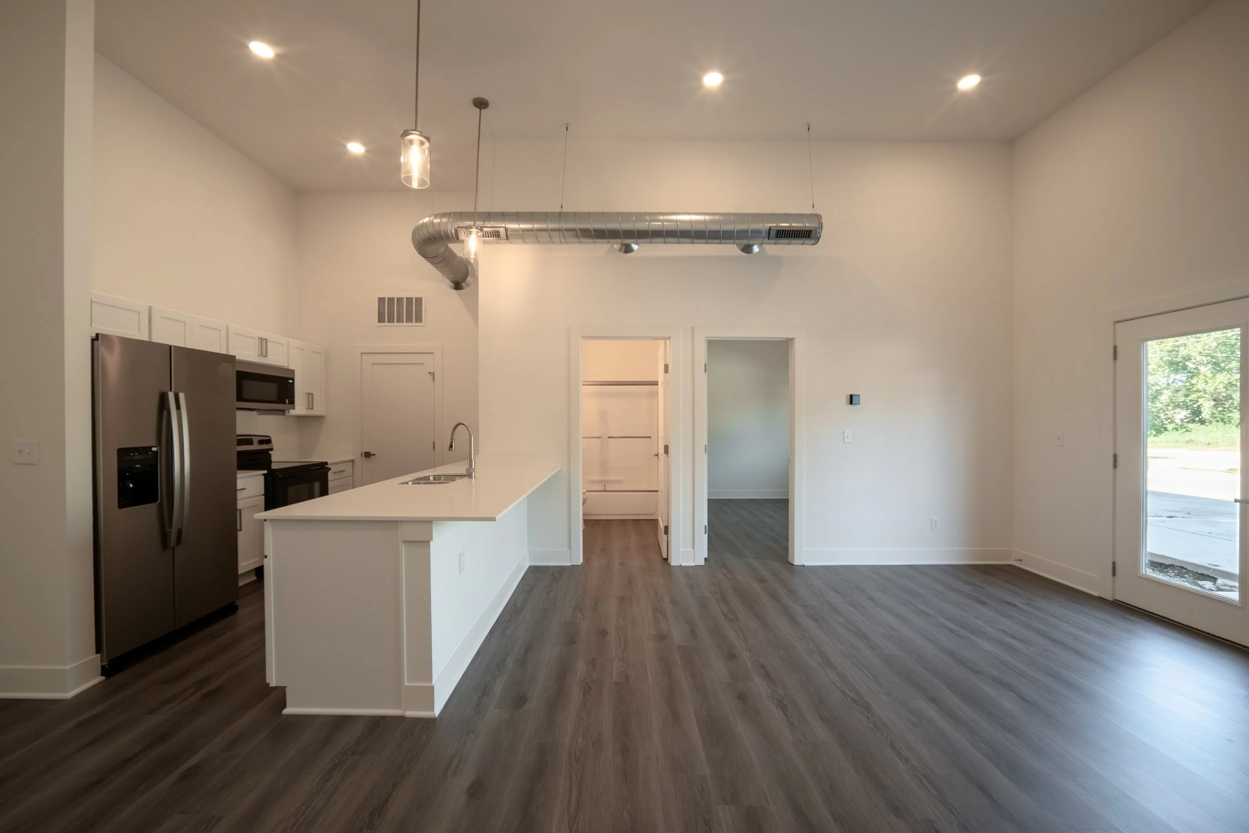 Interior of a modern, empty kitchen and living space with white walls, wood flooring, a stainless steel refrigerator, microwave, and black stove. An open doorway leads to a small room, and a large glass door opens to a backyard with greenery.