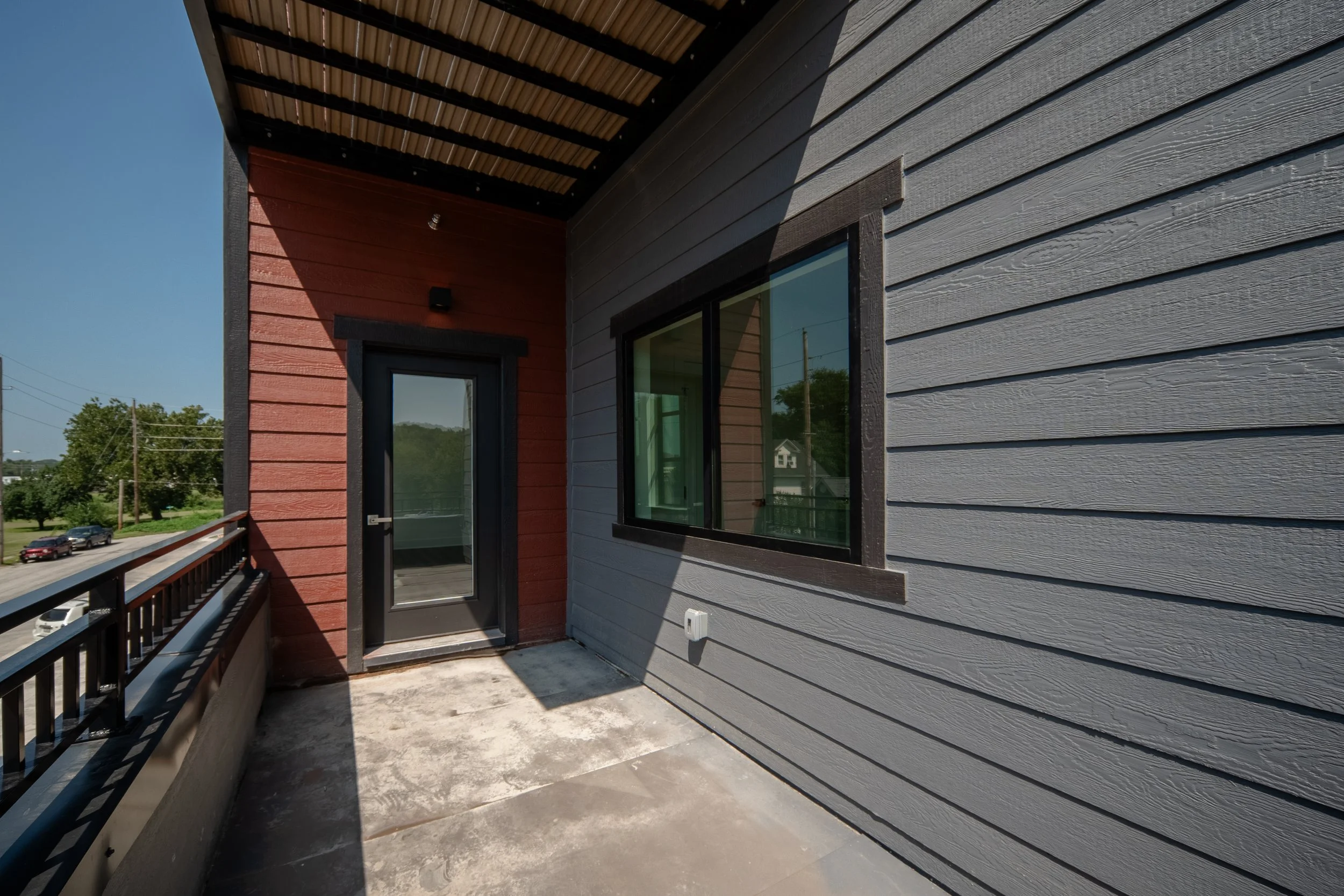 View from a balcony showing the exterior siding of a modern building with horizontal gray and red wooden planks, a door with glass panel, a window, and a metal railing, with a parking lot and greenery in the background.