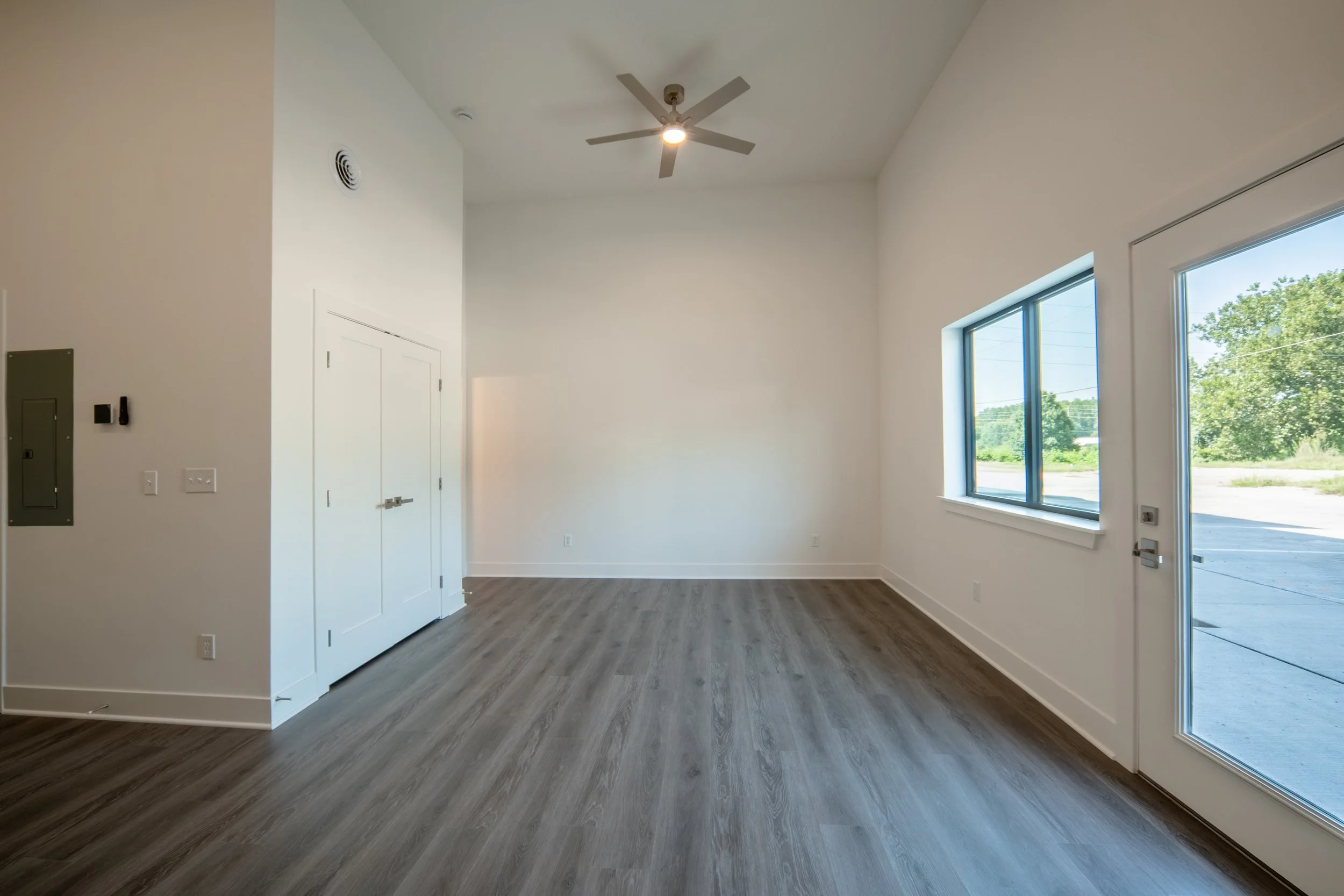 Empty room with large window, wood flooring, white walls, closet doors and a ceiling fan.