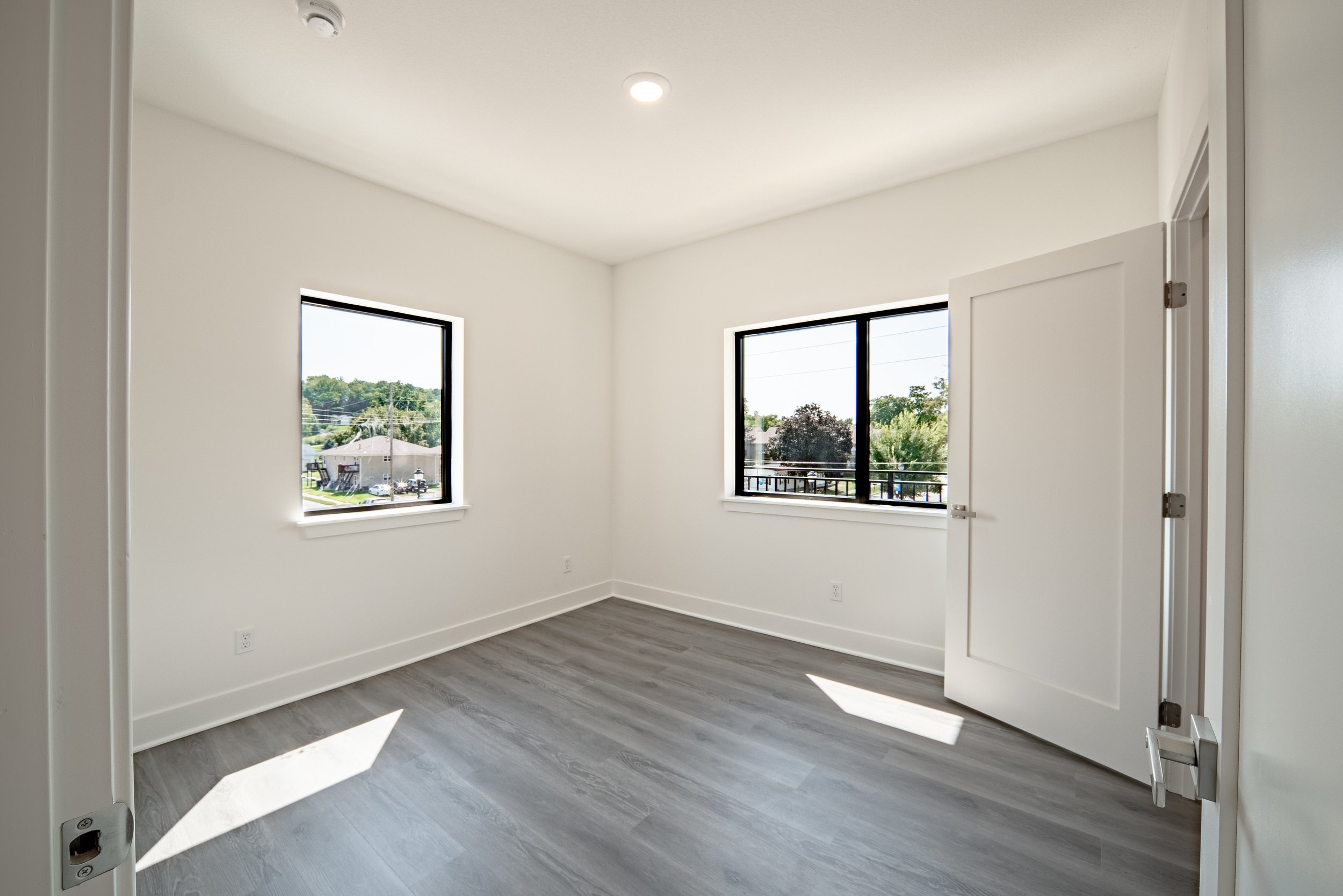 Empty room with white walls, two large windows showing a suburban neighborhood view, and a partially open door.