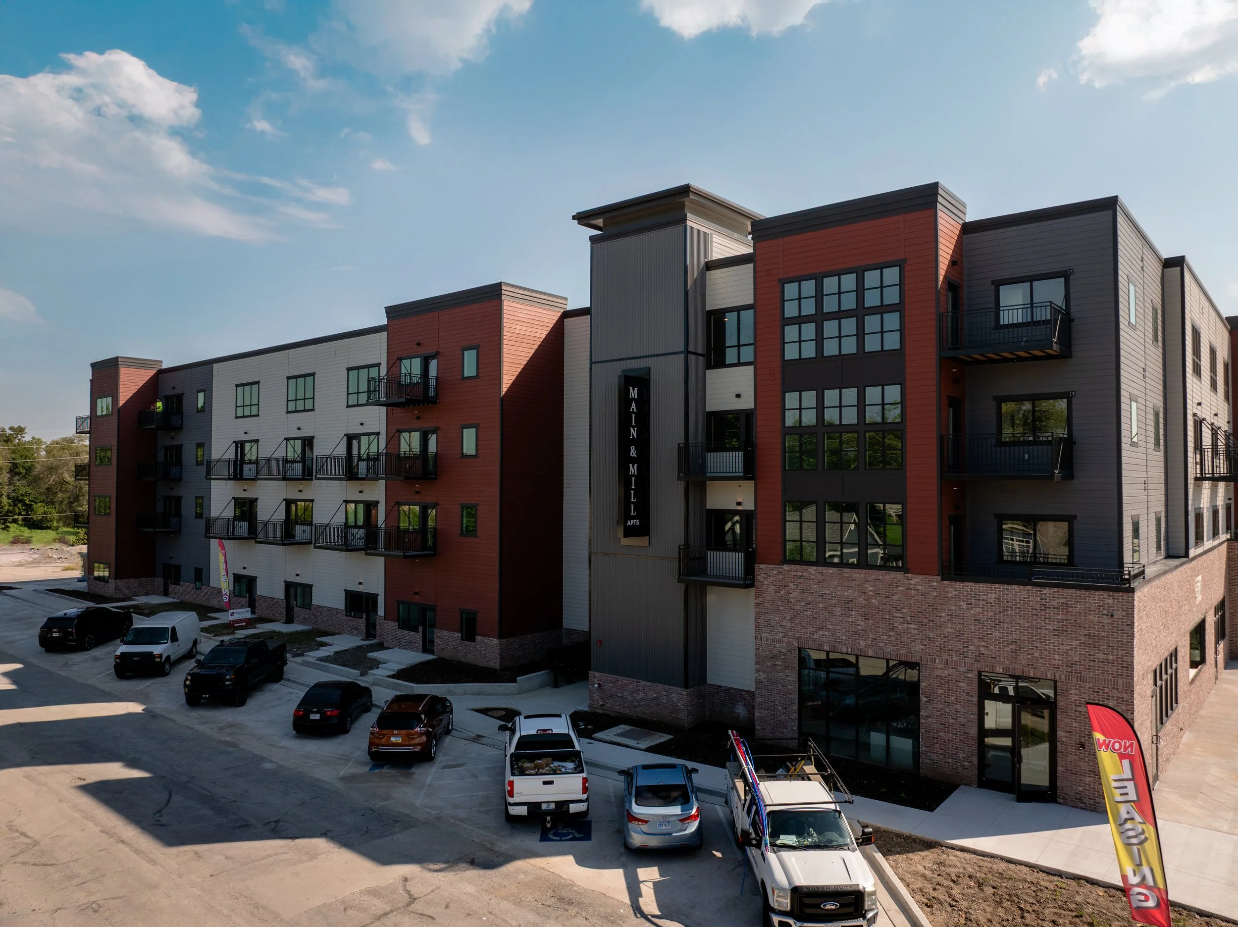 Modern, multi-story apartment building with a mix of brick, gray, black, and red exterior panels. Multiple balconies with black railings and large windows. Plenty of parking in front. Clear sky with some clouds.