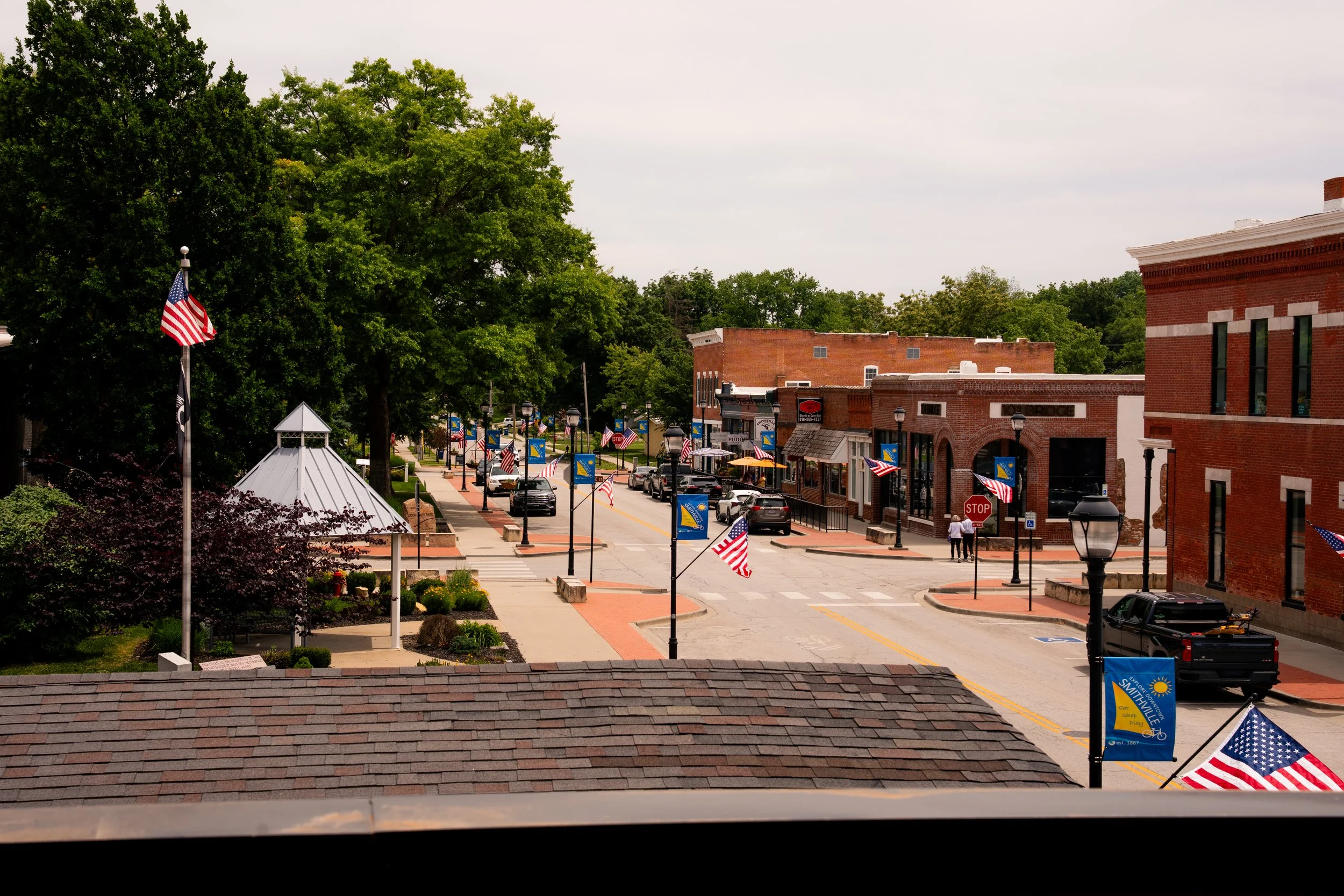 A small town main street decorated with American flags and banners, lined with brick buildings and lush green trees under a cloudy sky.