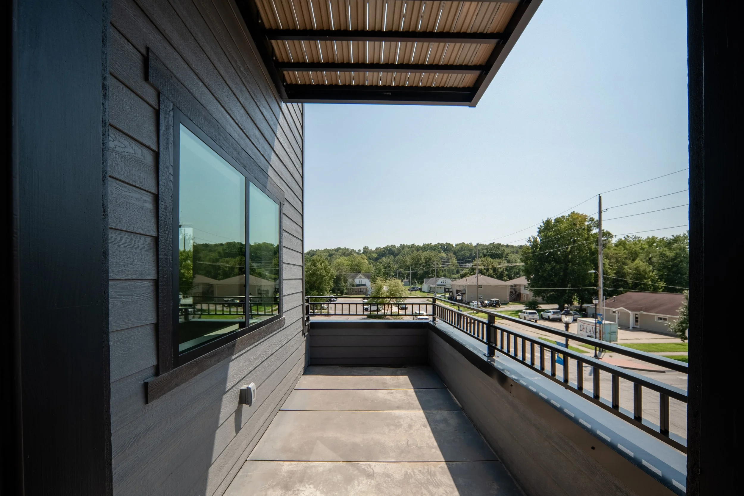 View from a balcony showing residential neighborhood with houses, trees, and utility poles in the distance. The balcony has a wooden ceiling, dark gray siding, large window, and metal railing.