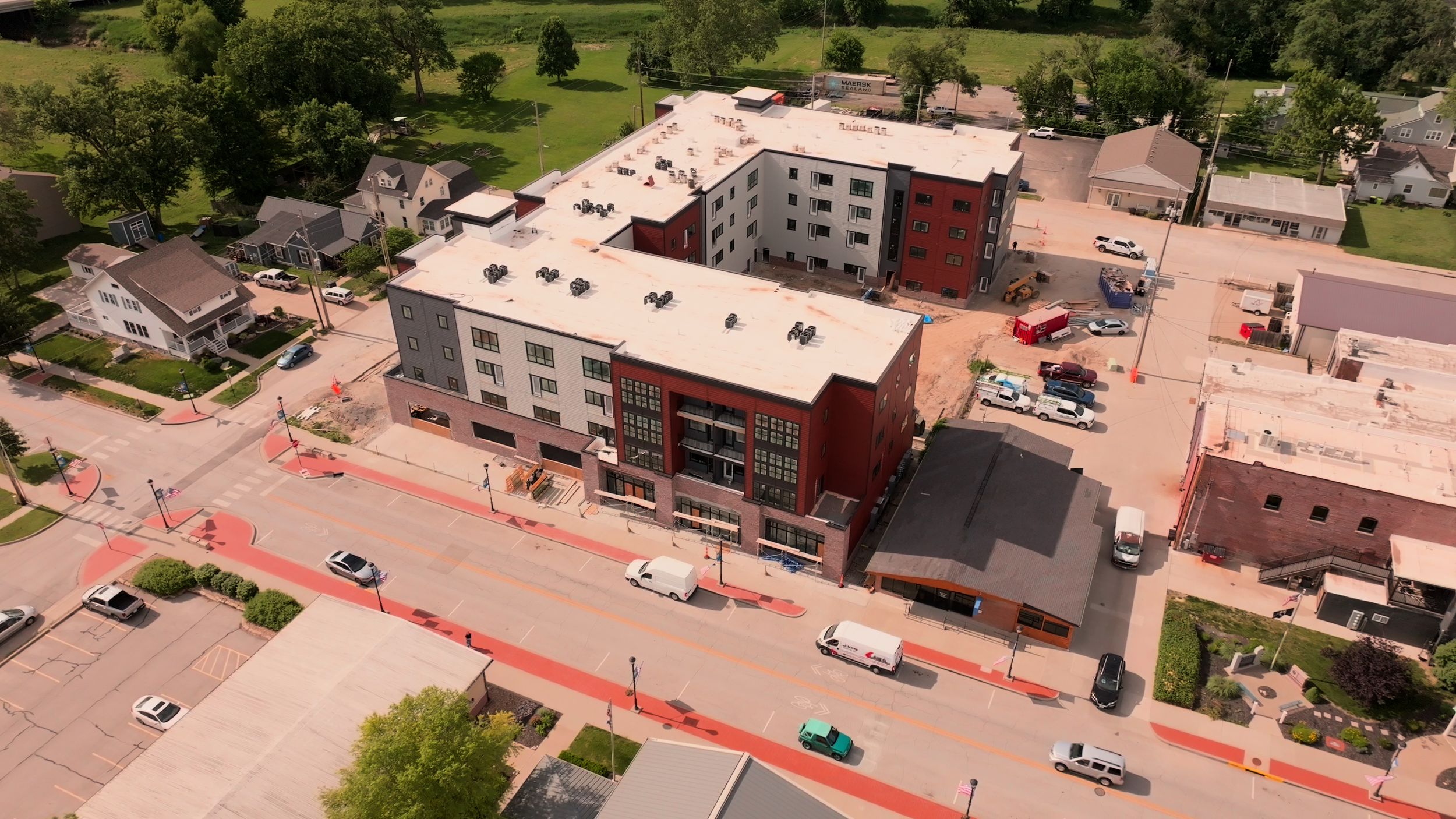 Aerial view of a city street next to a construction site with partly built multi-story buildings, parked cars, and surrounding green trees and residential houses.