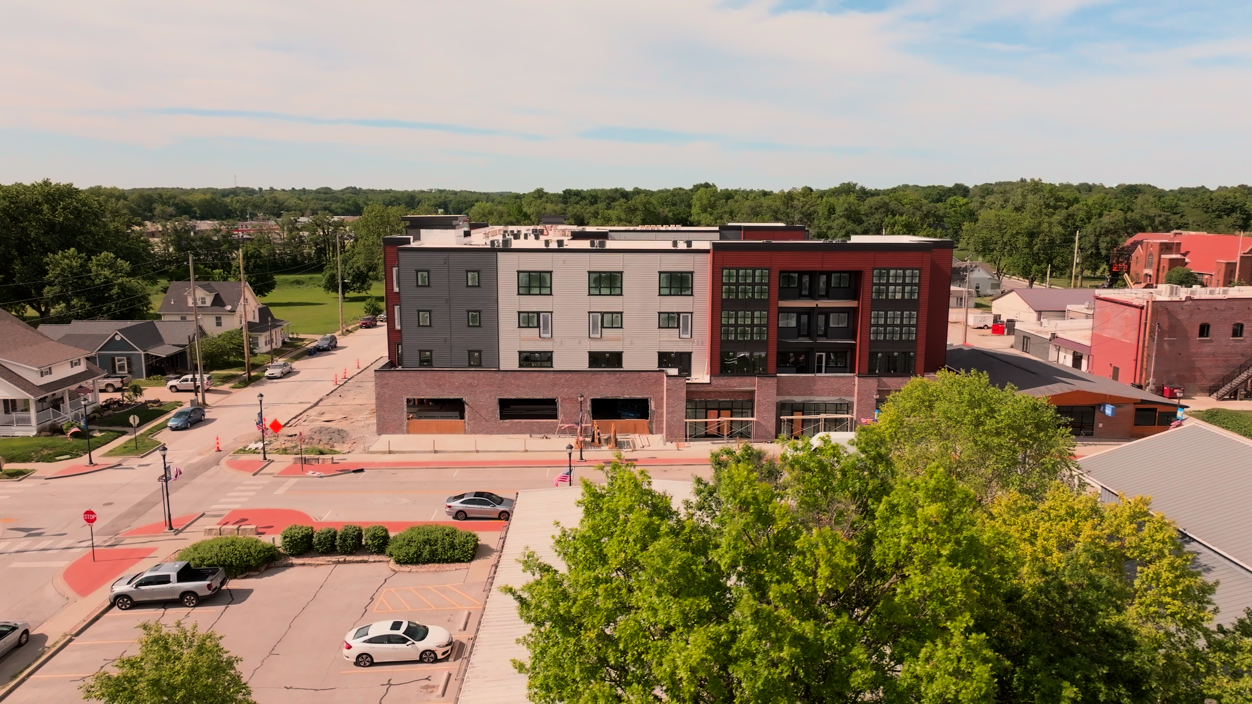 Aerial view of an apartment complex, a parking lot with several cars, trees, and a green landscape in the background under a partly cloudy sky.
