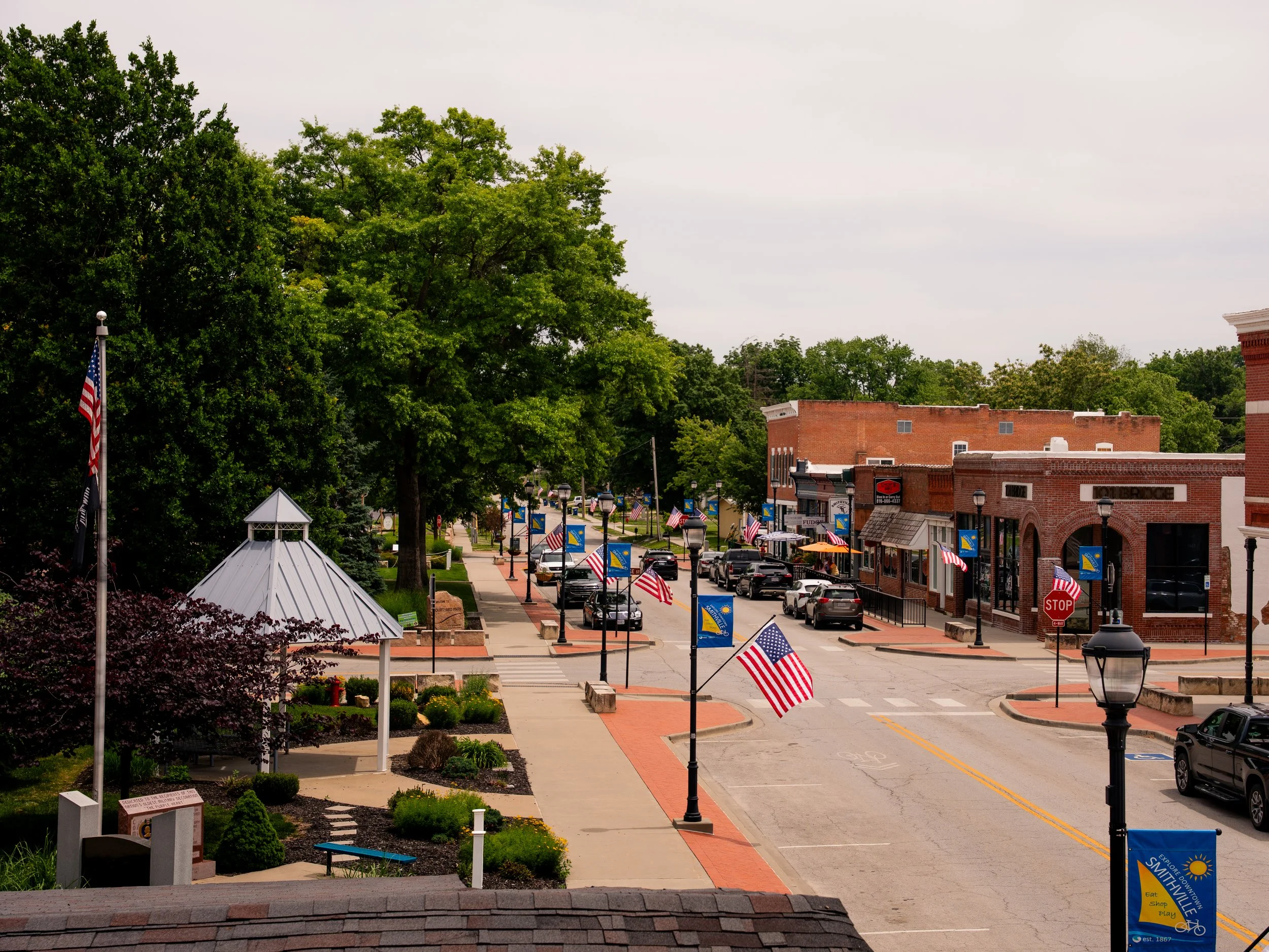 A small town street decorated with American flags and banners, lined with trees, brick buildings, and small outdoor shops, during daytime with cloudy sky.