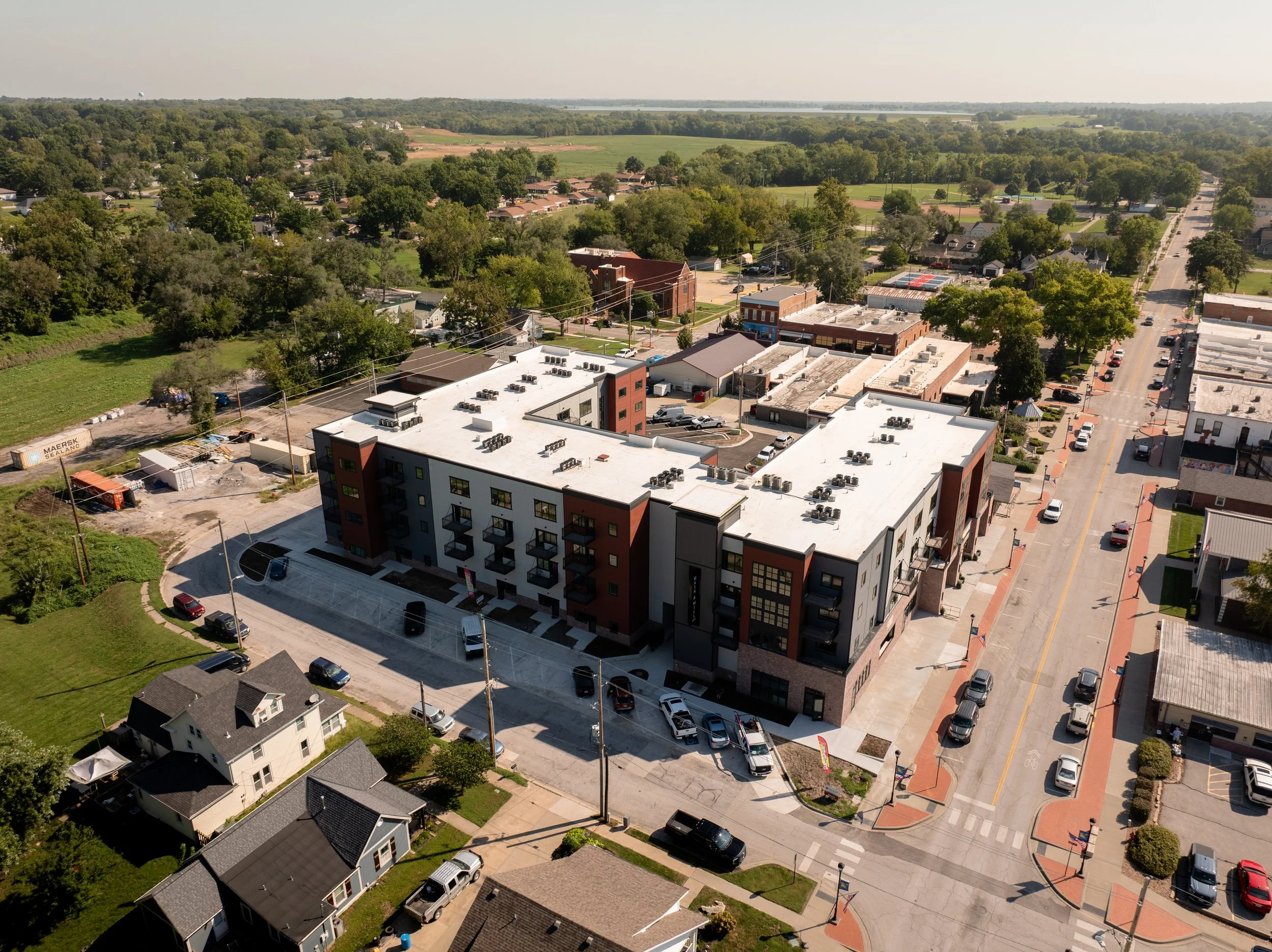 Aerial view of a small town with residential and commercial buildings, streets, trees, and surrounding green landscape with fields and Smithville lake in the distance.