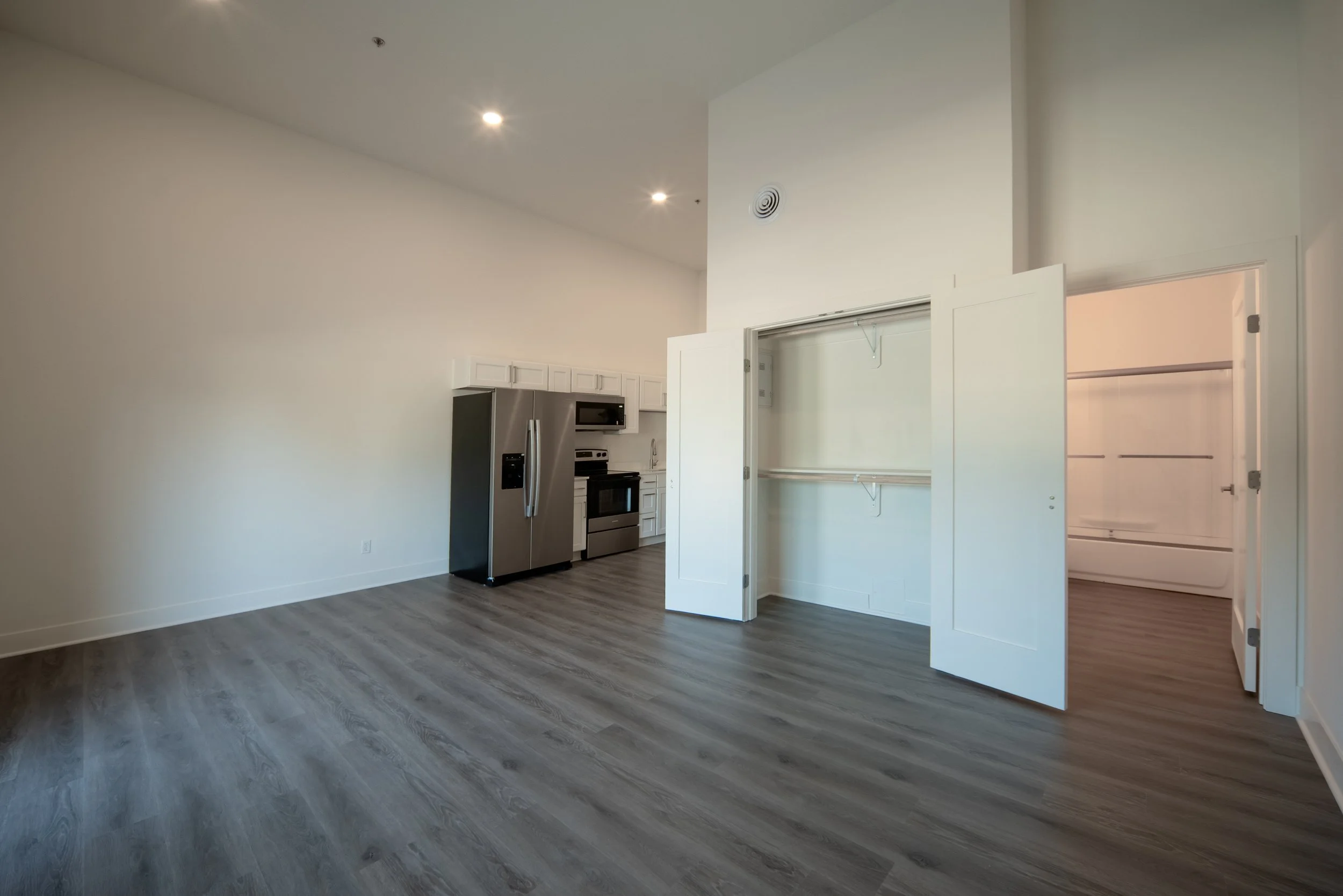 Open kitchen area with white cabinets, stainless steel refrigerator, microwave, and oven. Empty closet space with shelves in the foreground. Light-colored walls and wood flooring. Open studio living and bedroom space.