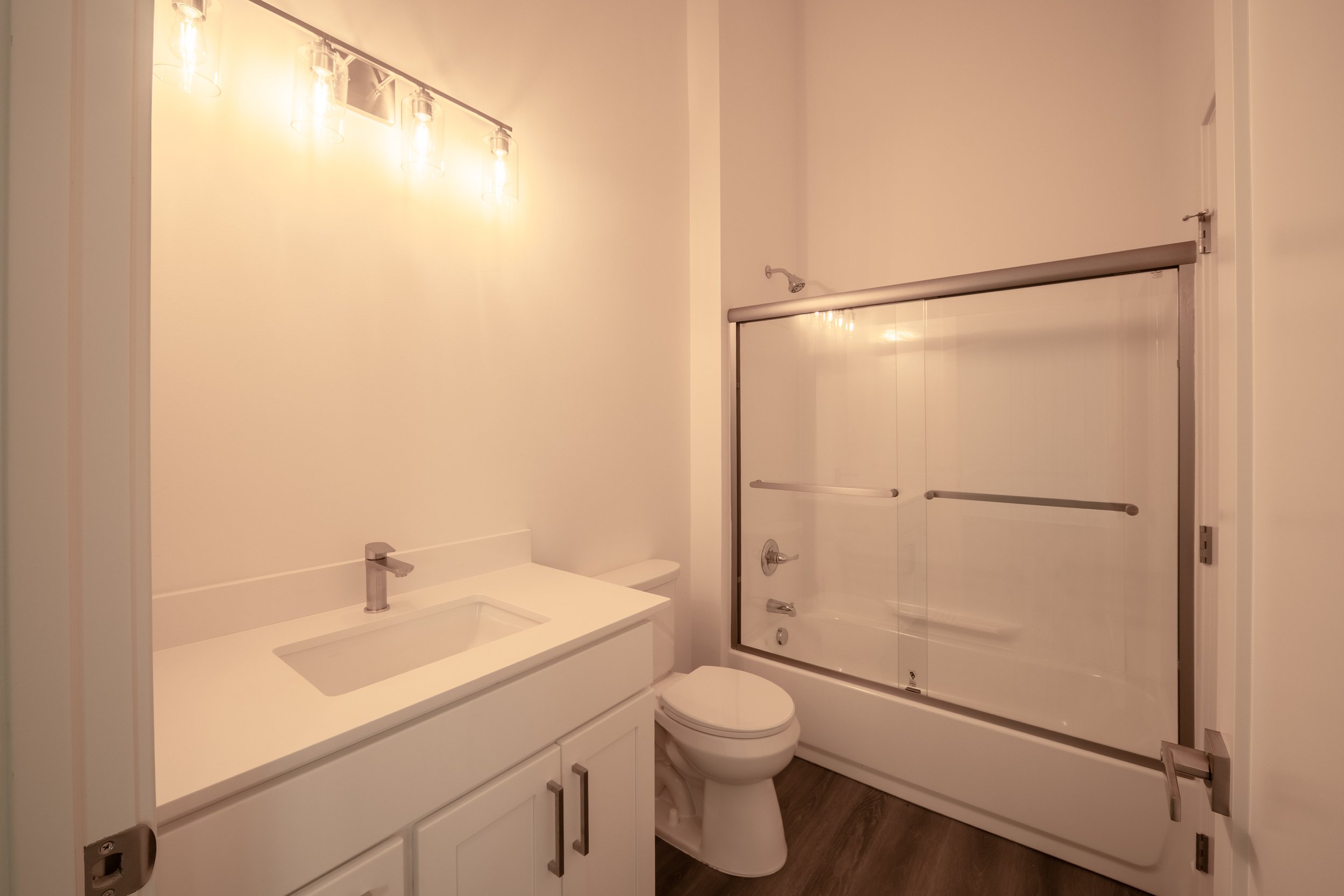 Modern bathroom with white vanity with storage, a sink with a chrome faucet, a toilet, and a glass shower with a sliding door. The bathroom has light-colored walls and dark wood flooring, illuminated by a row of three light fixtures.