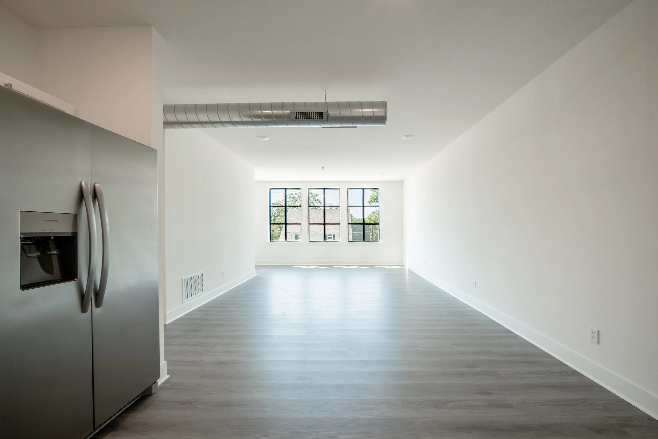 Empty modern apartment with large windows, white walls, gray flooring, and a stainless steel refrigerator.