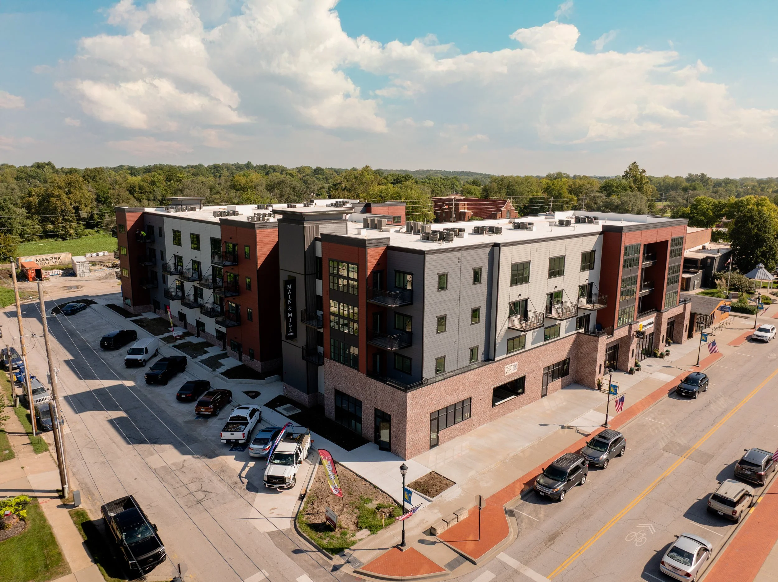 Aerial view of a modern multi-story apartment building with a parking lot, surrounded by trees and a blue sky with clouds. Restaurant located below apartments.