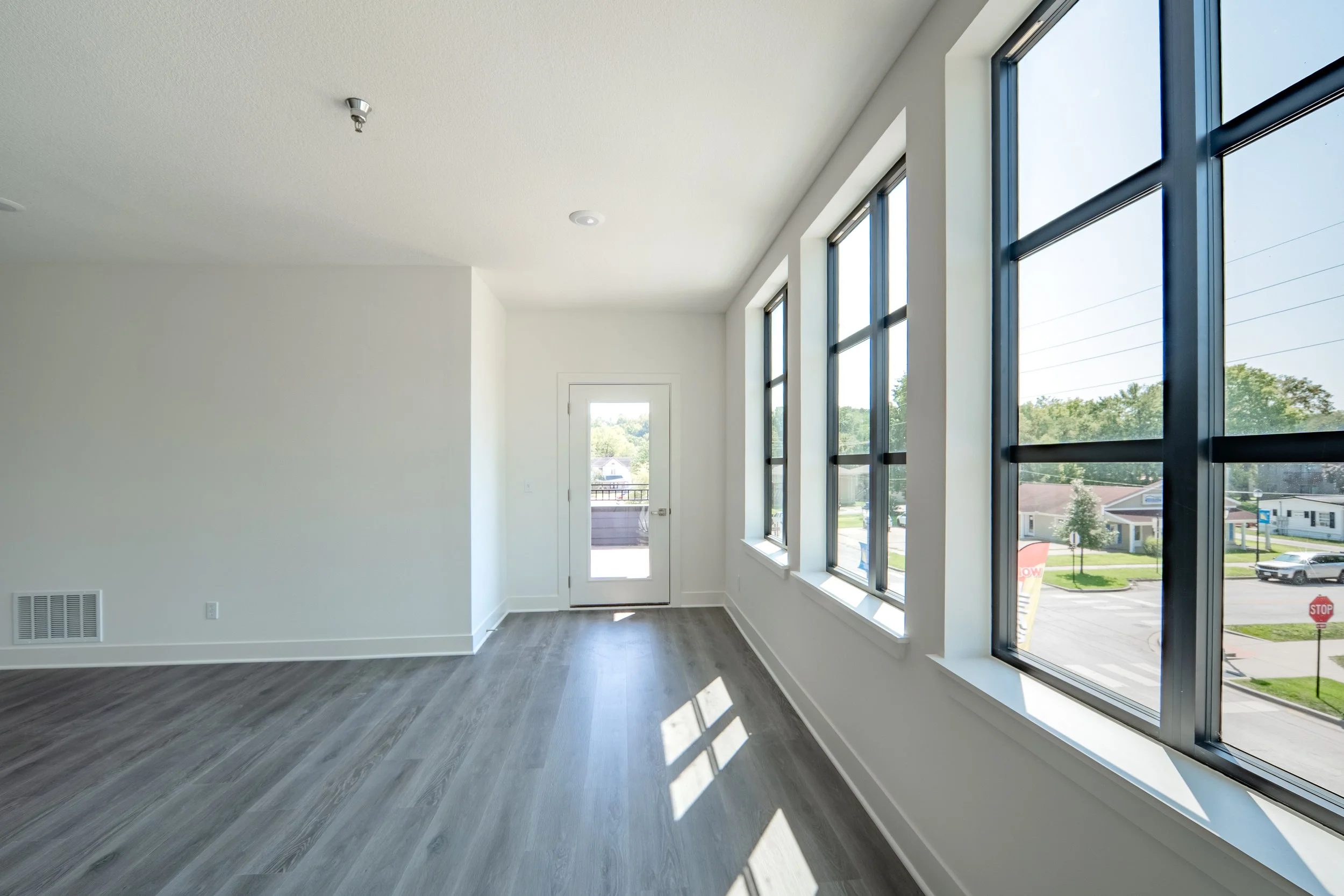 Empty room with large windows, hardwood flooring, white walls, and a white door leading outside.