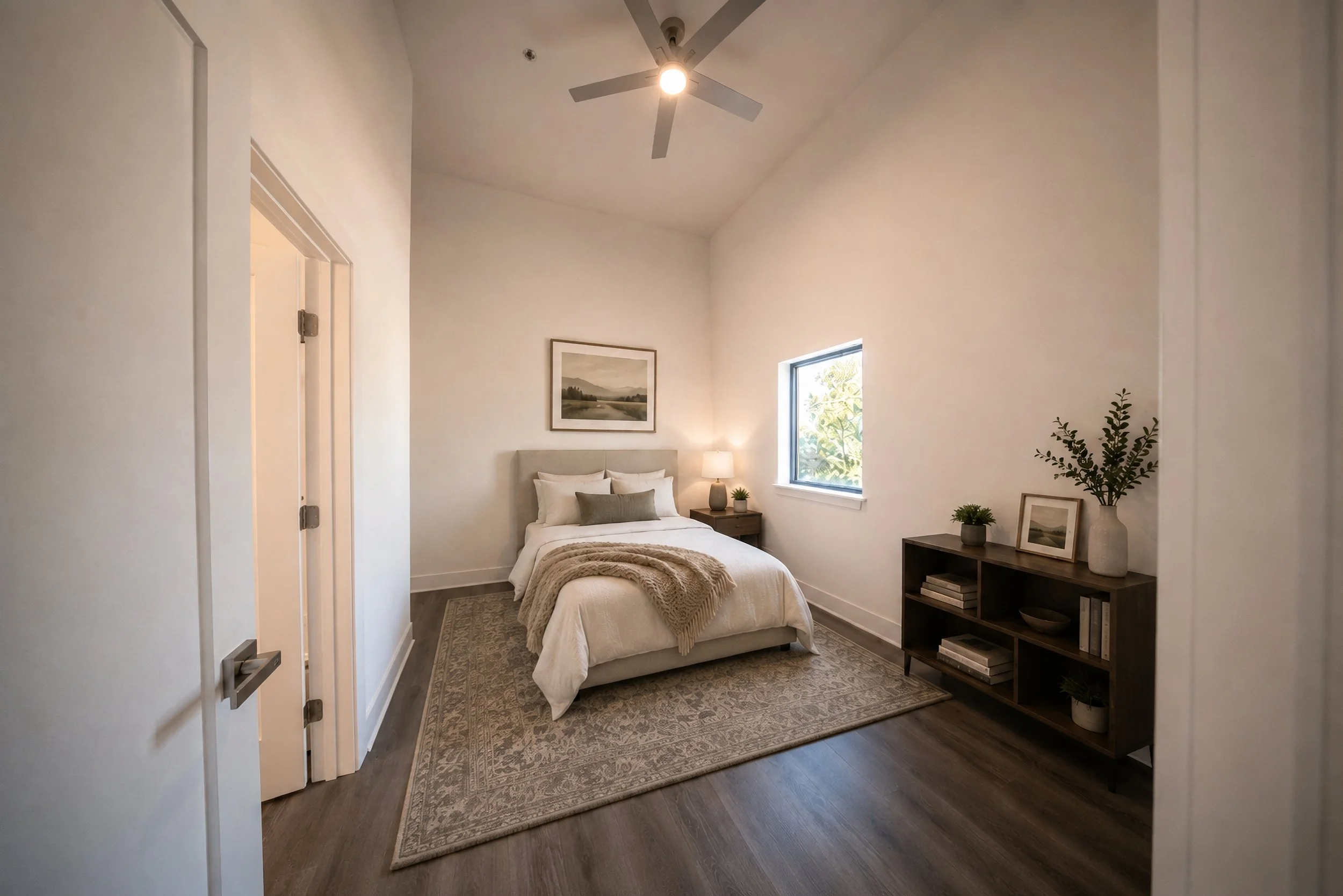 Virtually staged cozy bedroom with a large bed, beige headboard, and white bedding, decorated with pillows and a throw blanket. There is a nightstand with a lamp and small plants, a framed picture on the wall, an area rug, and a ceiling fan.
