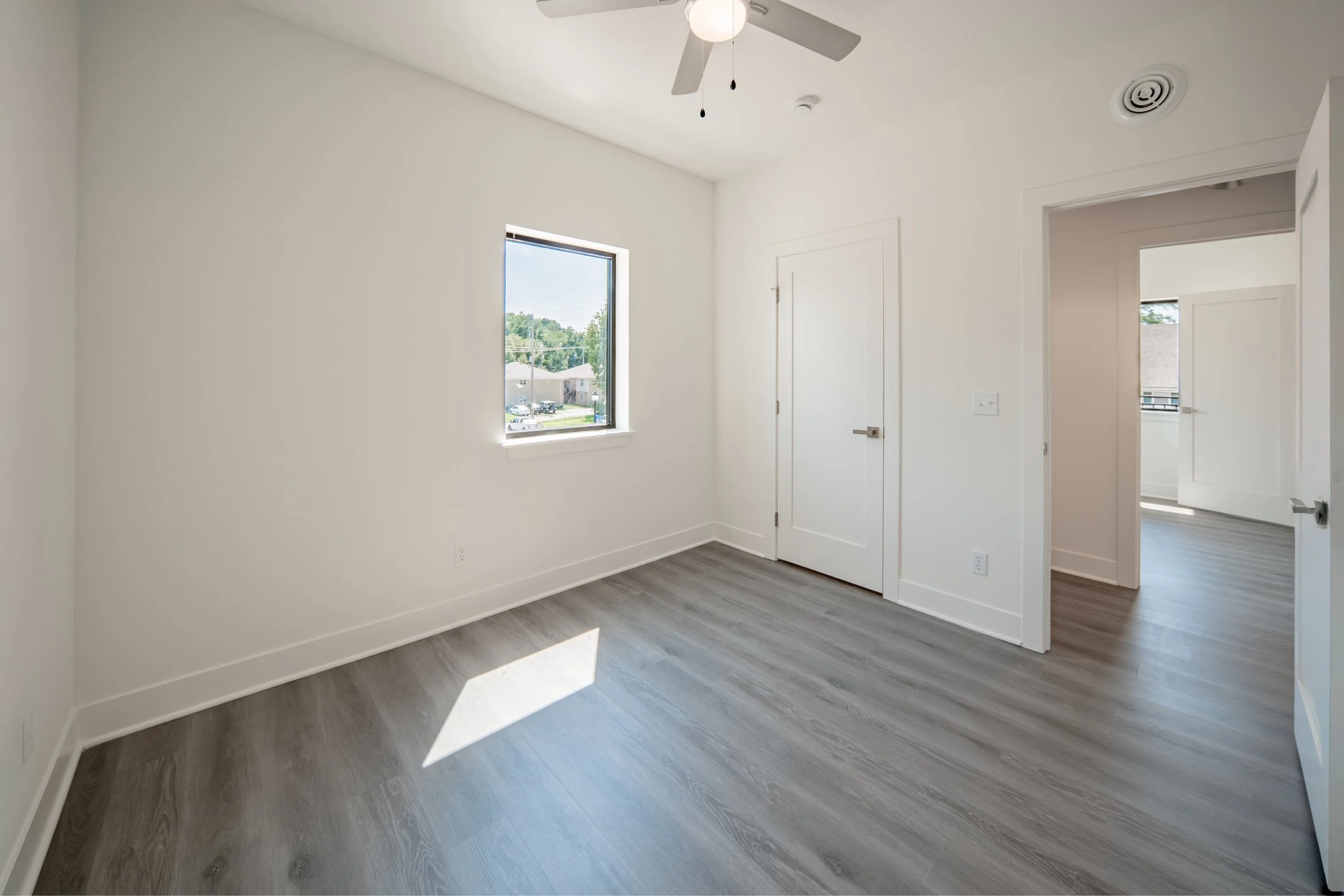 Empty room with white walls, gray wood flooring, a window showing a suburban neighborhood, a ceiling fan, and doors leading to separate areas.