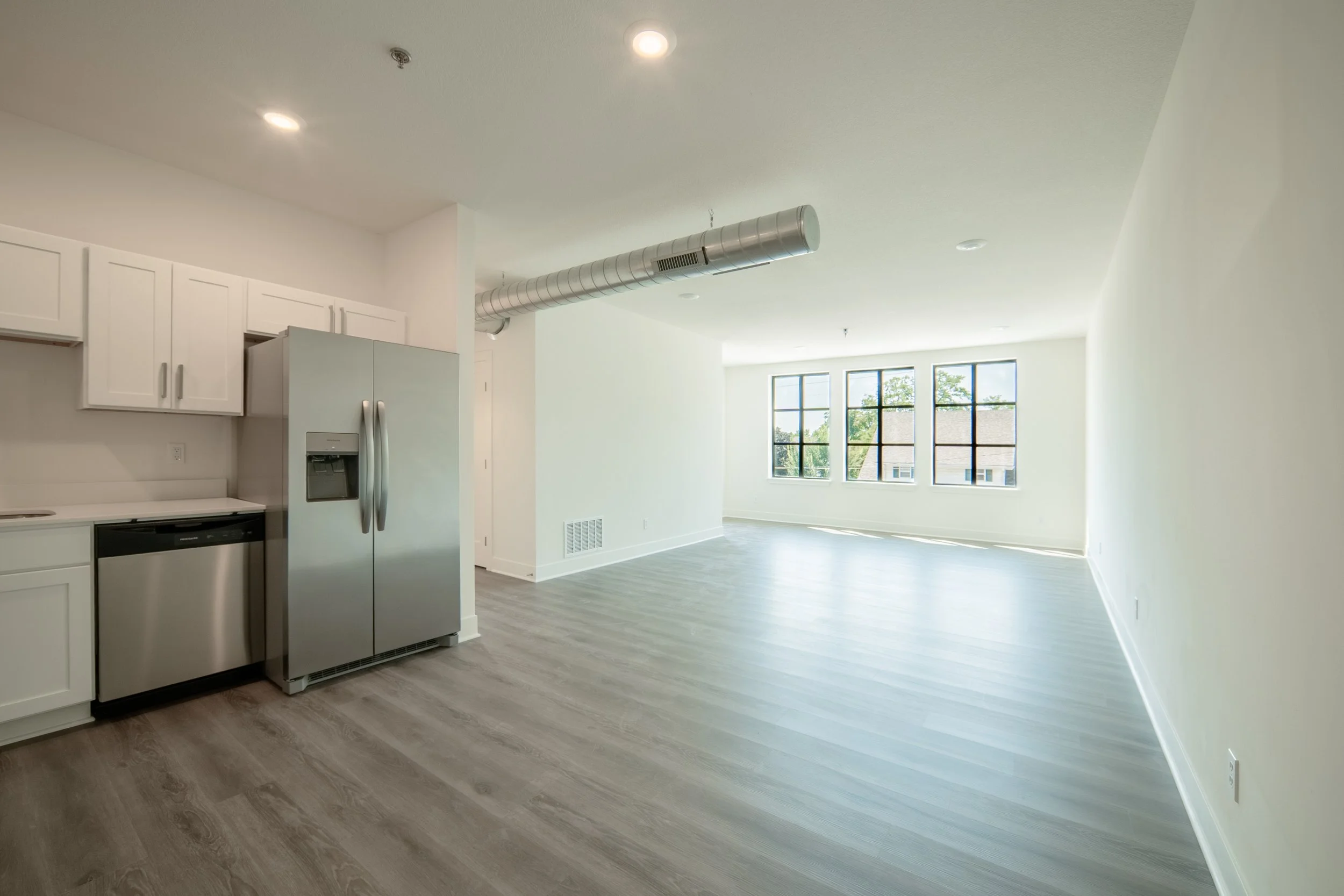 Open-concept apartment with white kitchen cabinets, stainless steel refrigerator, and large windows letting in natural light. Gray wood-look flooring throughout.