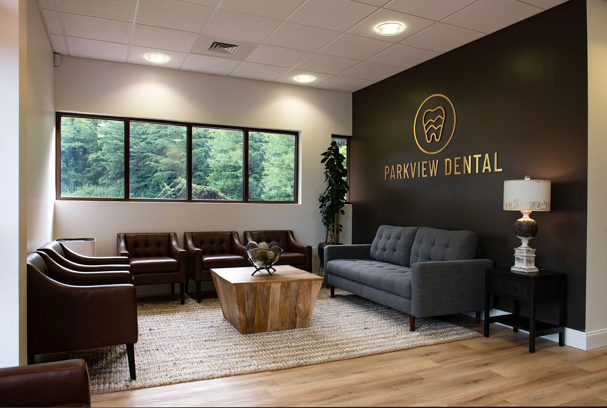 Parkview Dental Asheville office waiting room with cozy couches, a woven rug, a wooden coffee table, windows looking out at trees, and a gold logo large on a black wall